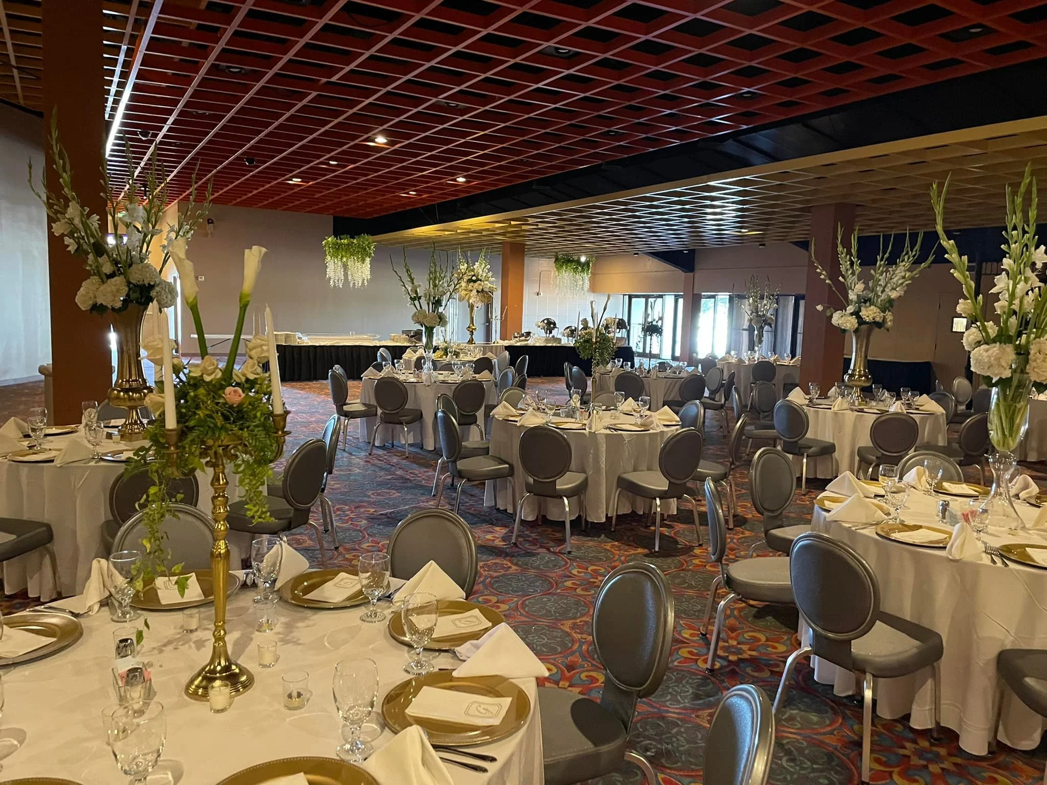 A banquet hall decorated with tall floral arrangements on round tables with white tablecloths, set with gold and white dinnerware, glassware, and napkins.