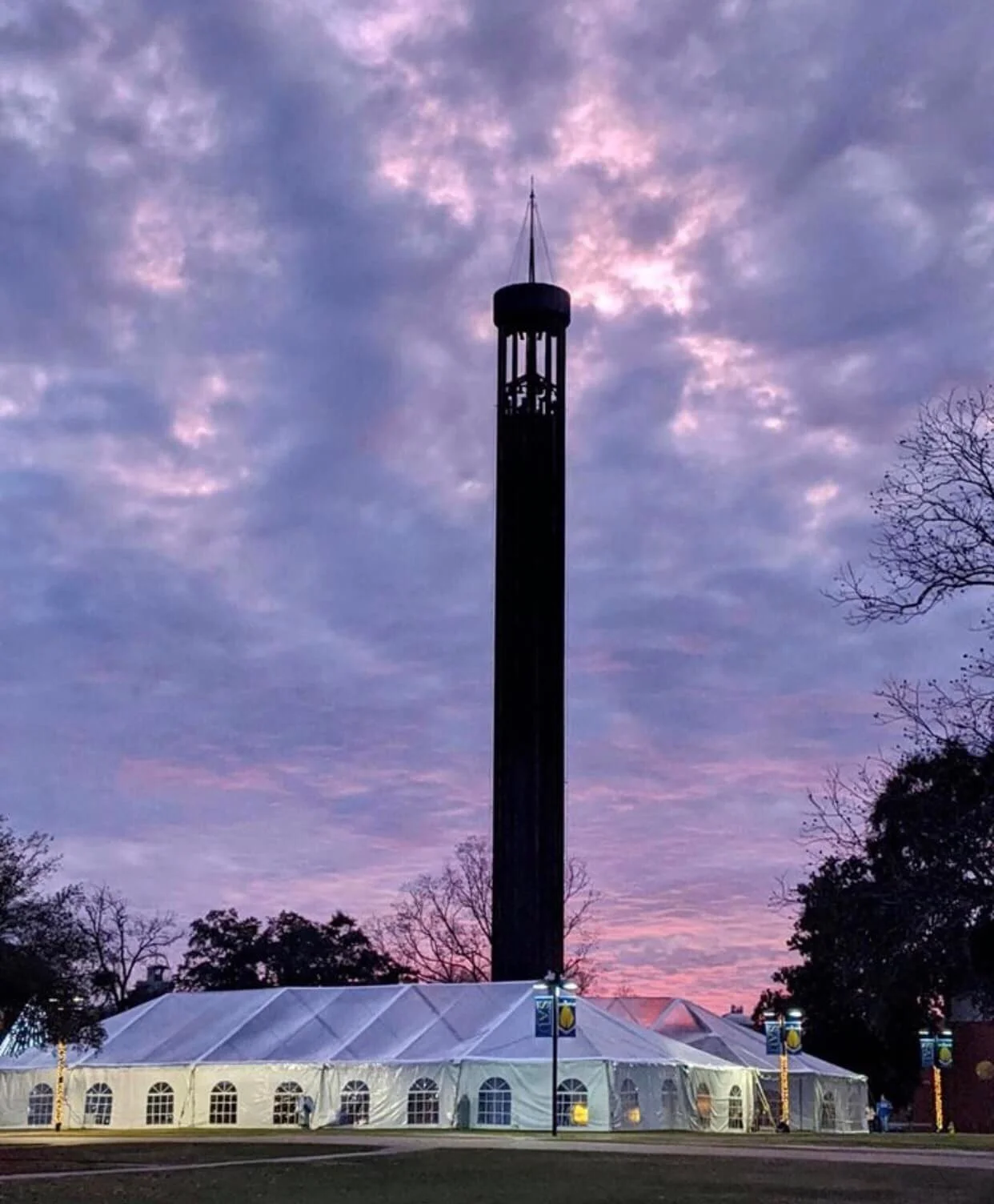 Tall black tower with a clock near the top, under a colorful evening sky with purple and pink clouds. There is a large white tent with arched windows at the base of the tower, and trees are visible on either side.
