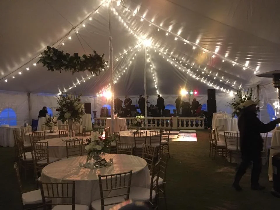 A decorated event tent with string lights, round tables with floral centerpieces, and a dance floor. There are people in the background near a stage with musicians and speakers, and some people are standing or walking inside the tent.