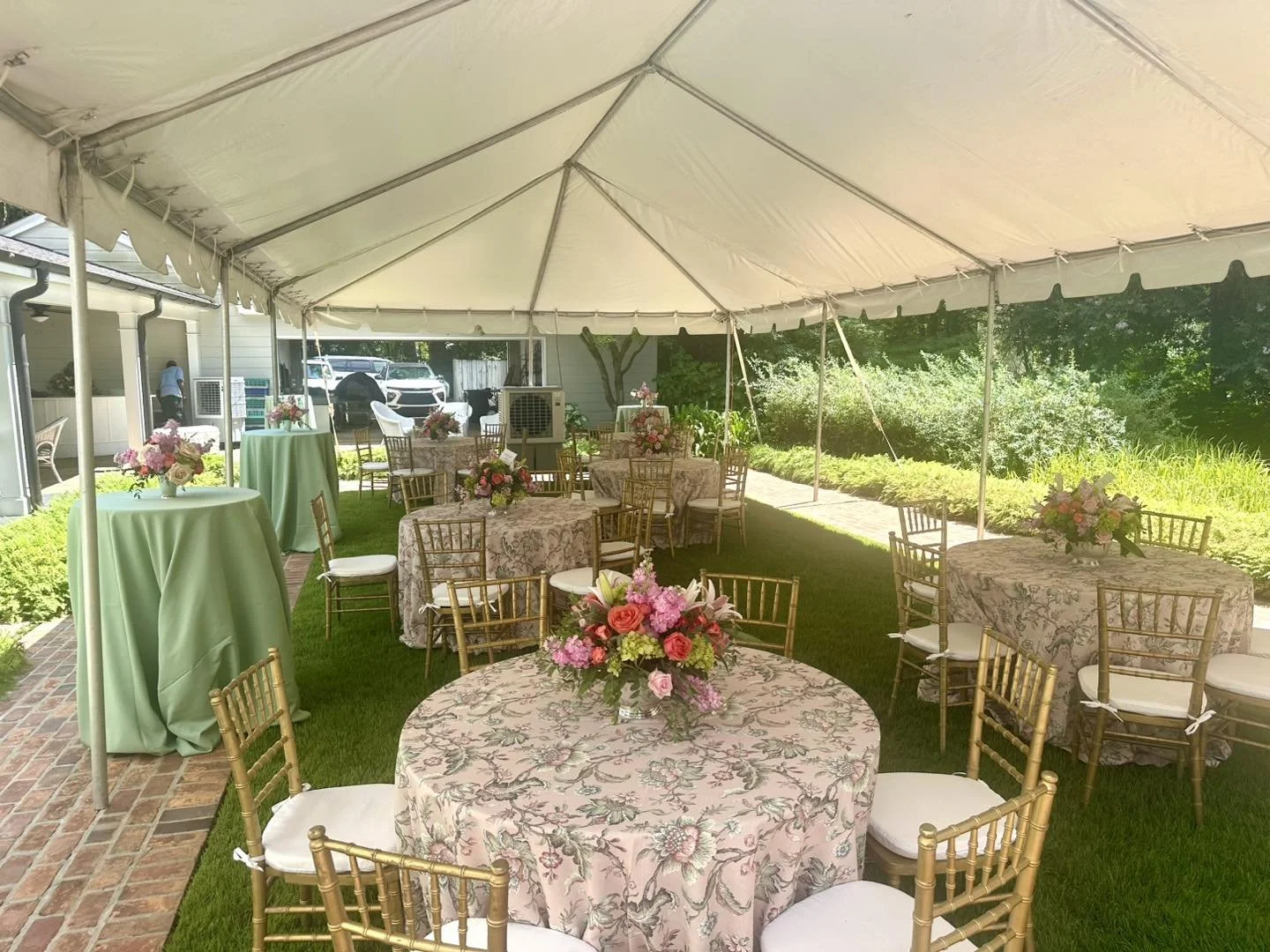Outdoor event setup under a large white tent with round tables covered in floral tablecloths, decorated with pink and red flower centerpieces, and surrounded by gold chairs with white cushions, in a garden setting.