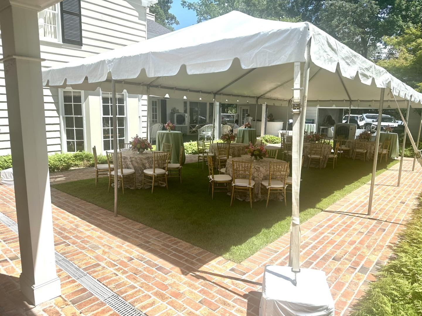 Outdoor event setup under a white canopy tent with round tables covered in floral tablecloths and flower centerpieces, surrounded by gold chairs, on a grassy area with brick pathway and greenery in the background.