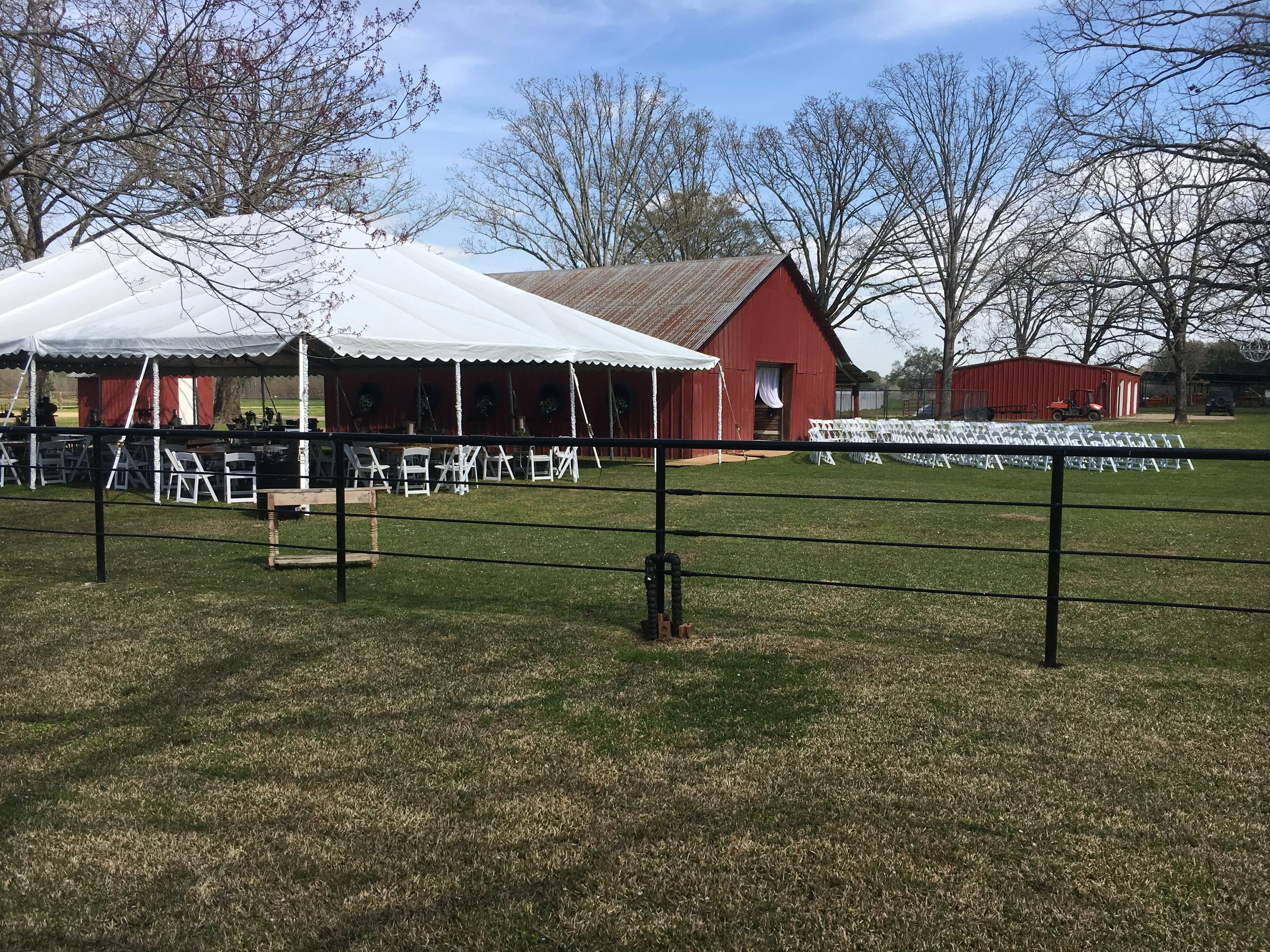 A large outdoor wedding setup with white chairs arranged in rows and a white tent, situated in front of a red barn with a rusted metal roof. Several leafless trees are in the background under a blue sky.