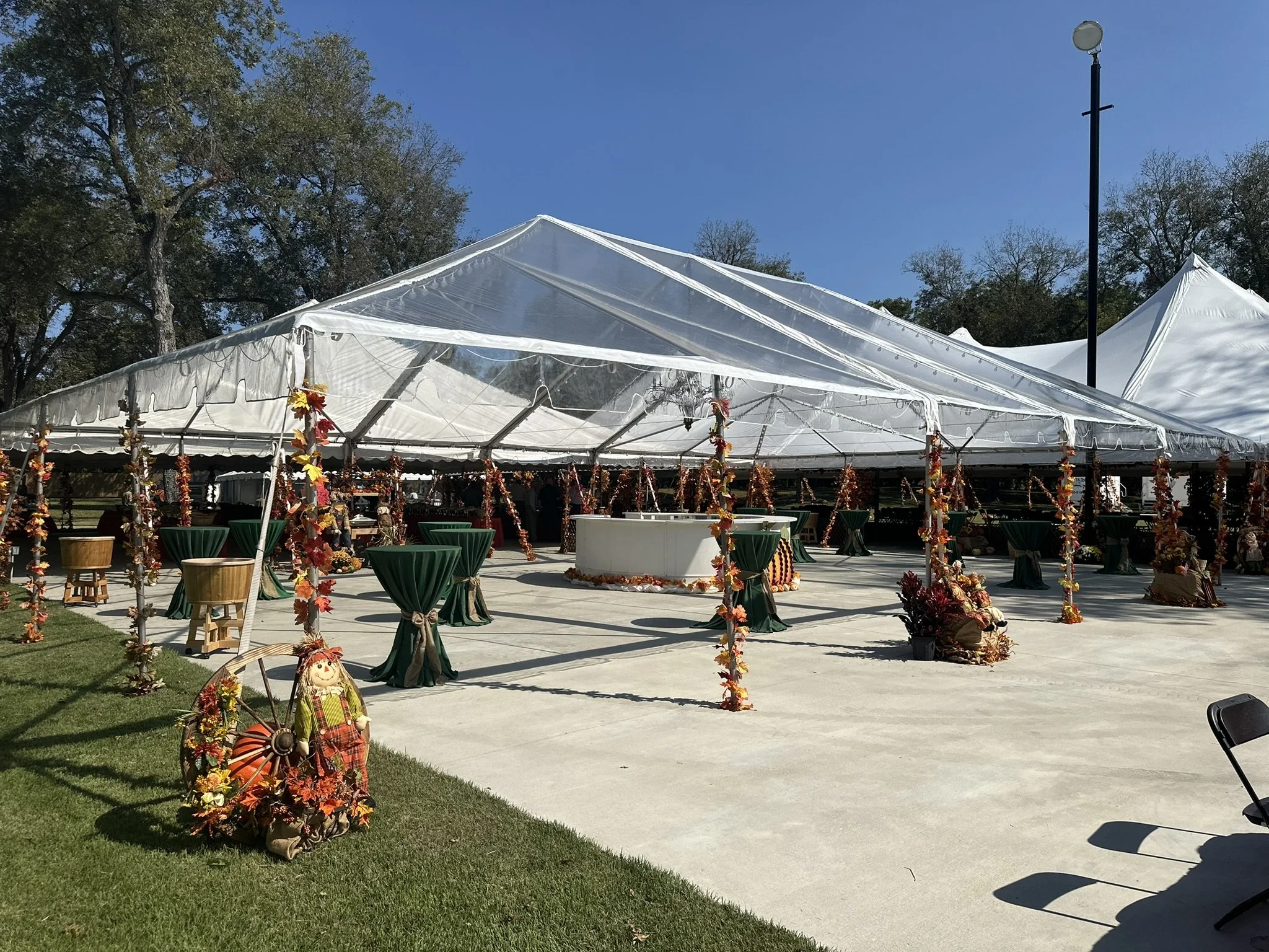 An outdoor event setup with a large white tent decorated with fall-themed garlands and a scarecrow figure on the ground nearby. There are tall tables draped with green cloths and a few chairs, with a grassy area and trees in the background under a cl