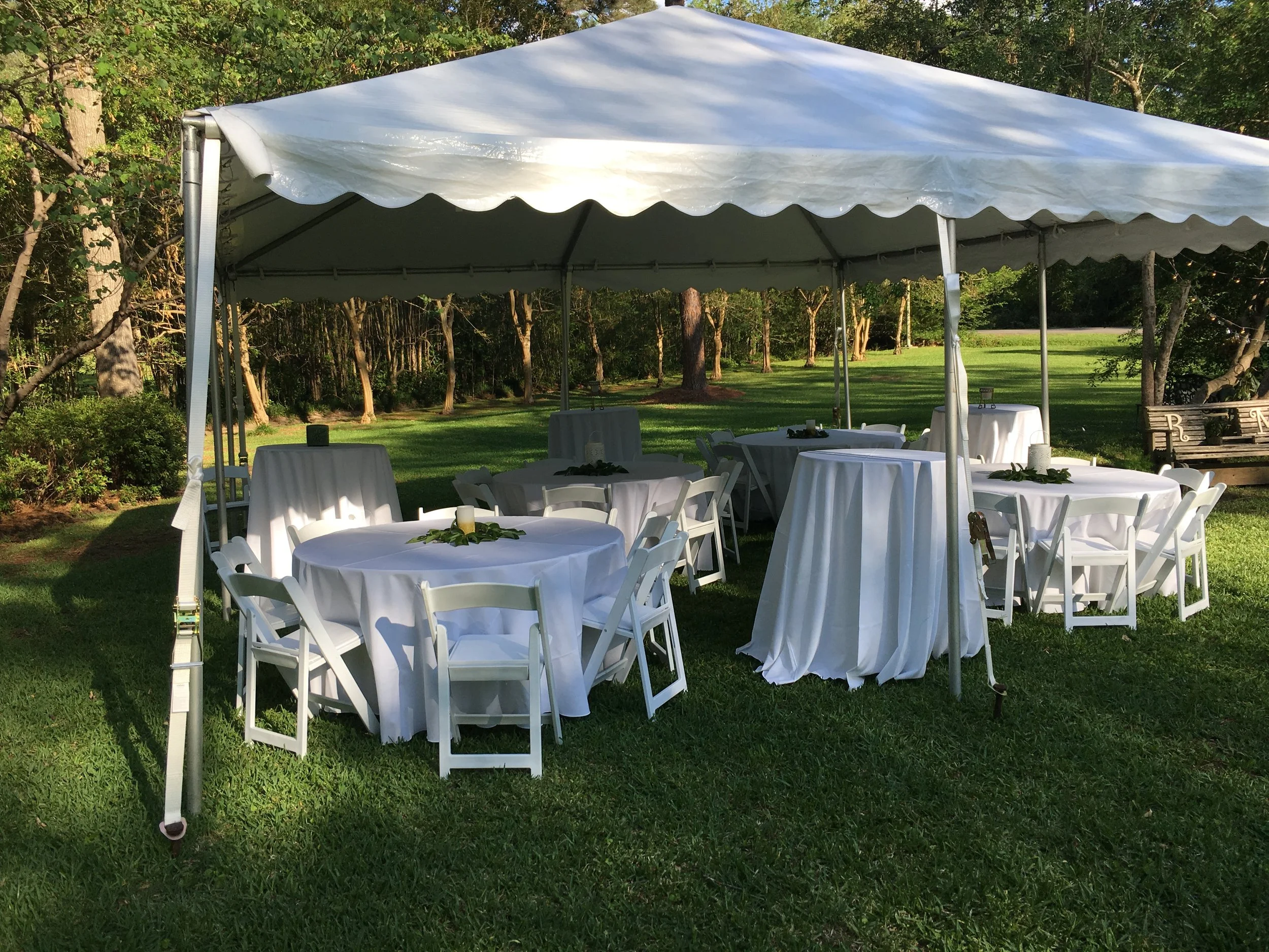 White outdoor event tent set up on a grassy lawn, with round tables covered with white tablecloths and surrounded by white chairs, in a park-like setting with trees in the background.