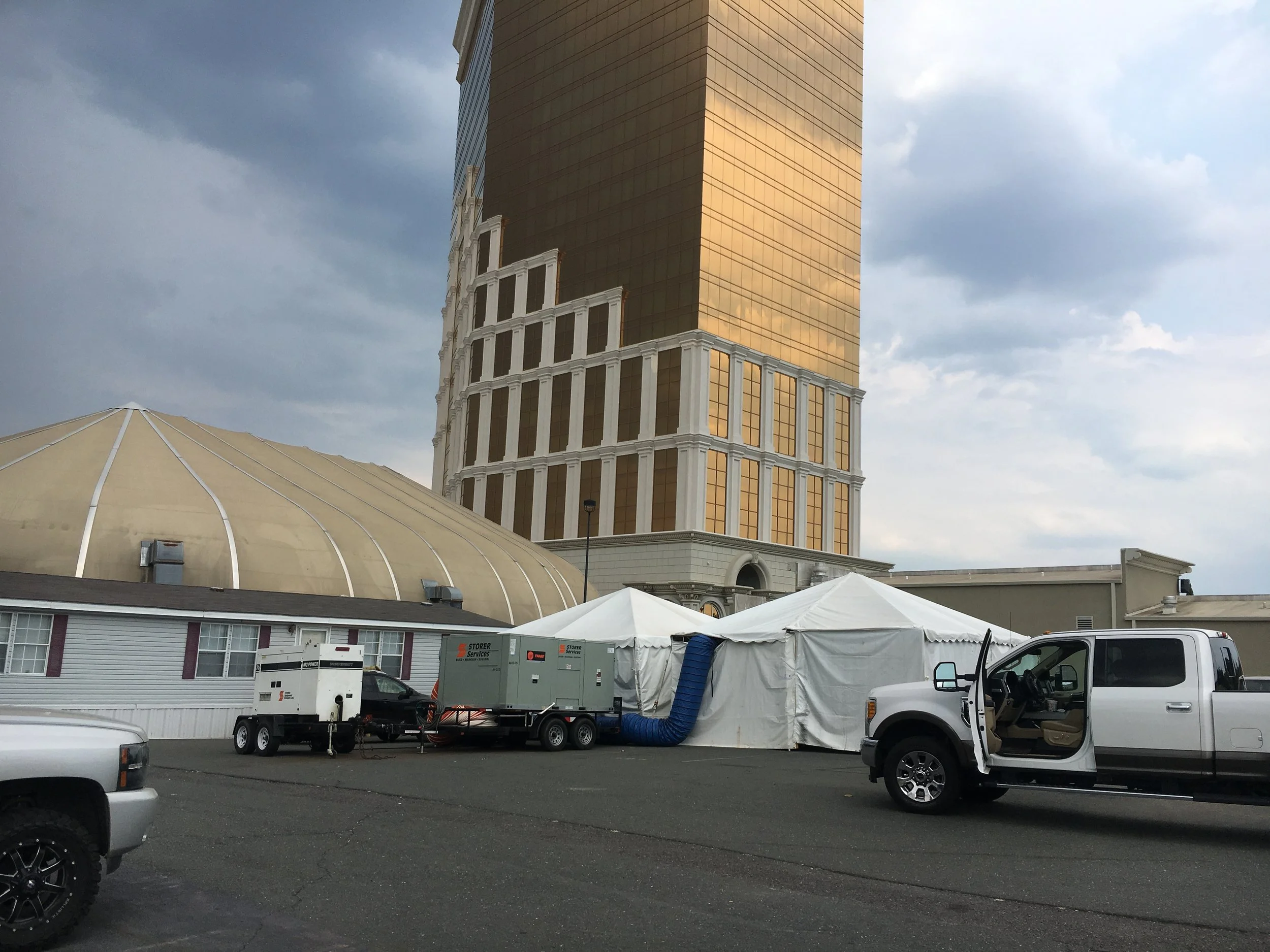 A construction or event setup area with a large white tent, smaller trailer, and vehicles in front of a tall, multi-story building with golden reflective windows. The sky is cloudy.