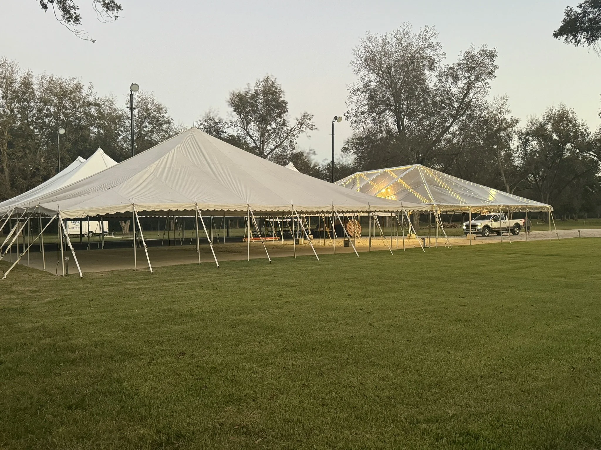 Two large white event tents set up on a grassy field, one with string lights, with trees and parked vehicles in the background during dusk.