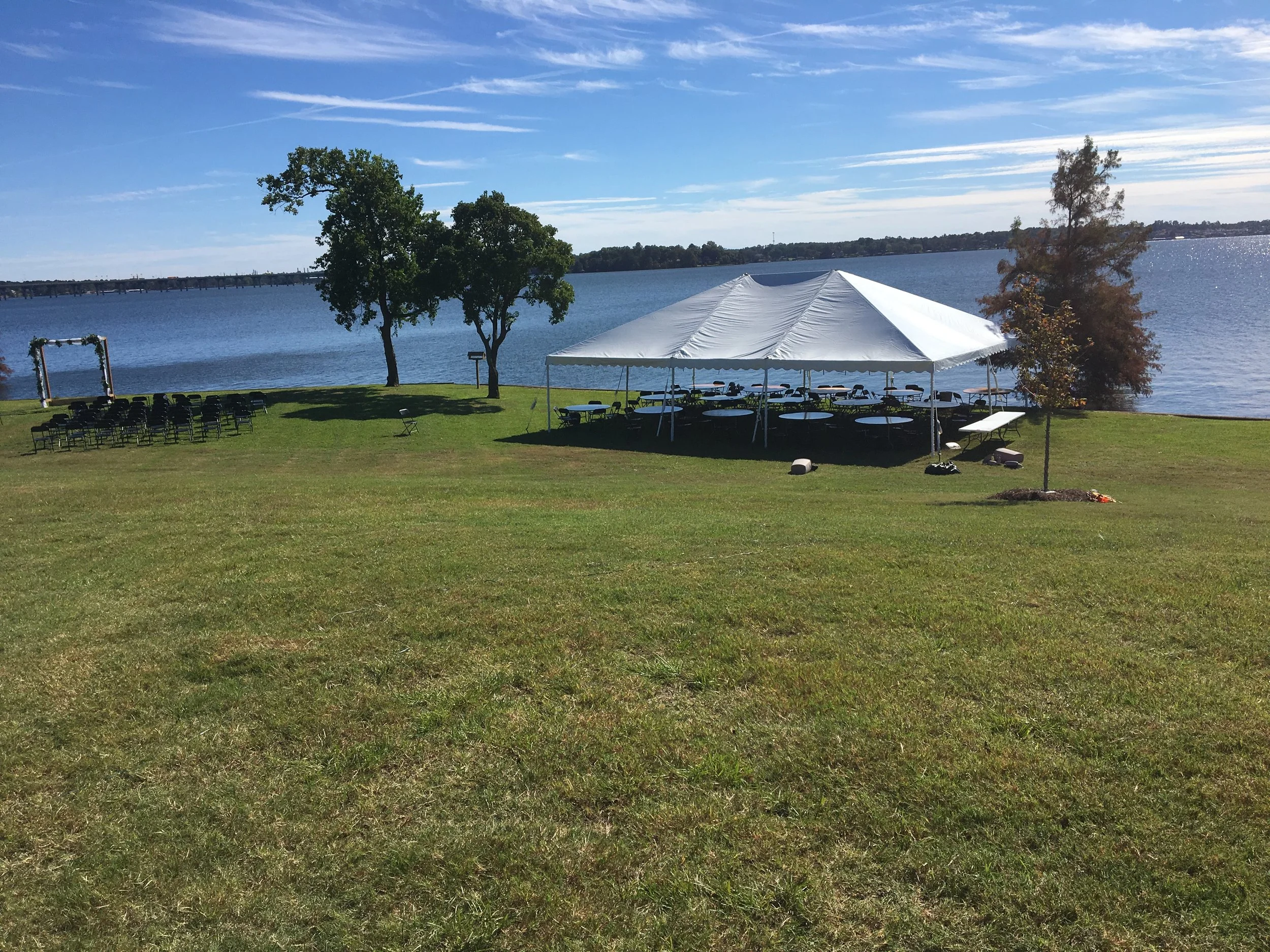 A lakeside outdoor event setup with a large white tent, chairs, and tables on a grassy area near the water, with trees and a clear blue sky in the background.