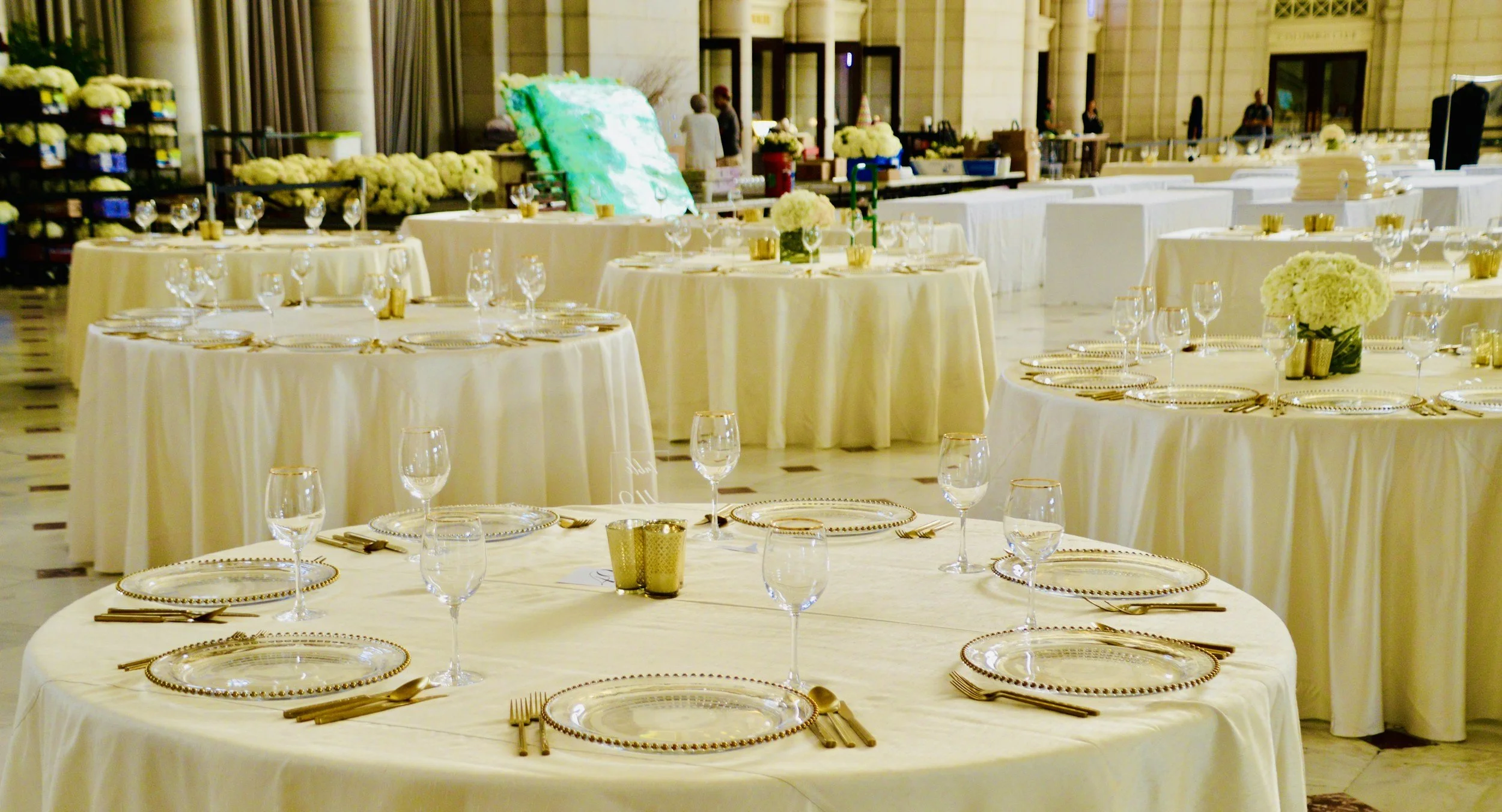Elegant banquet setup with round tables covered in cream tablecloths, adorned with floral centerpieces, glassware, gold utensils, and decorative plates in a spacious hall decorated for an event.