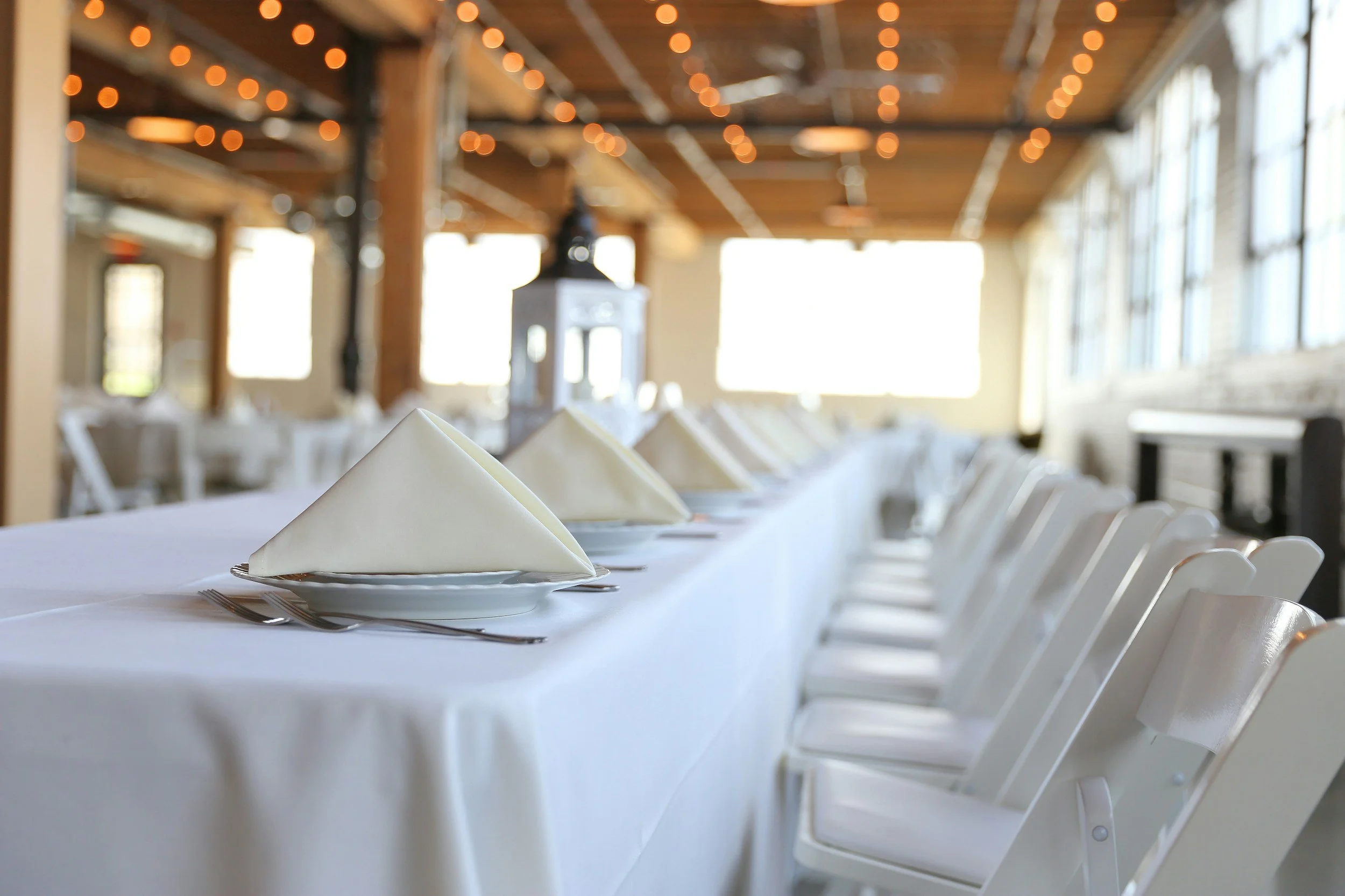 A long dining table with white tablecloths, neatly folded napkins on plates, and white chairs in a bright, rustic restaurant or event space with string lights and large windows.
