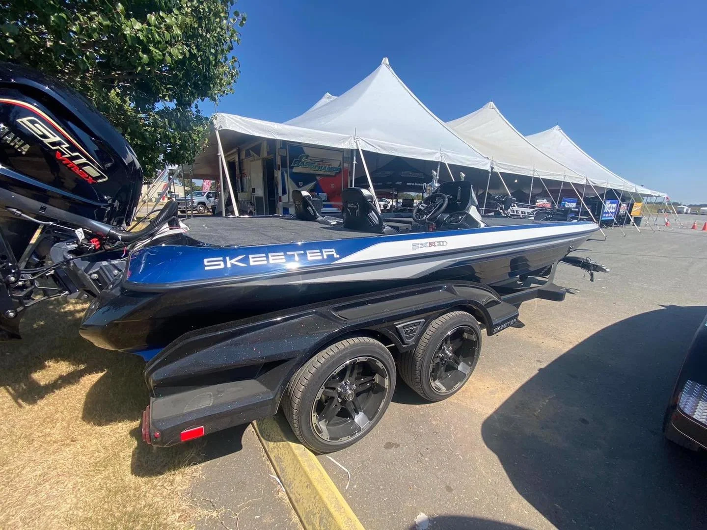 A black Skeeter fishing boat on a trailer with two seats and a Mercury outboard motor, parked outdoors under a blue sky near a large white event tent.
