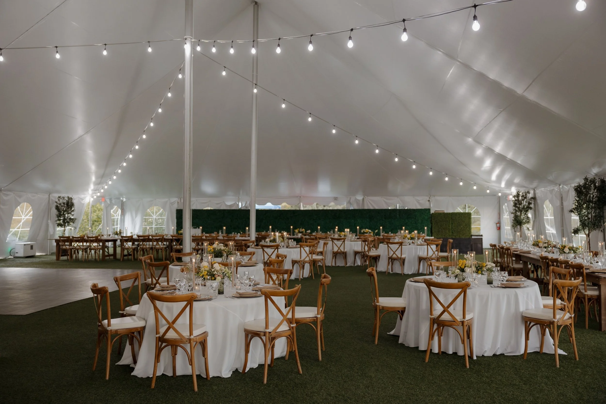 Elegant event tent decorated with string lights, tables with white tablecloths, floral centerpieces, and wooden chairs, set for a celebration or wedding reception.