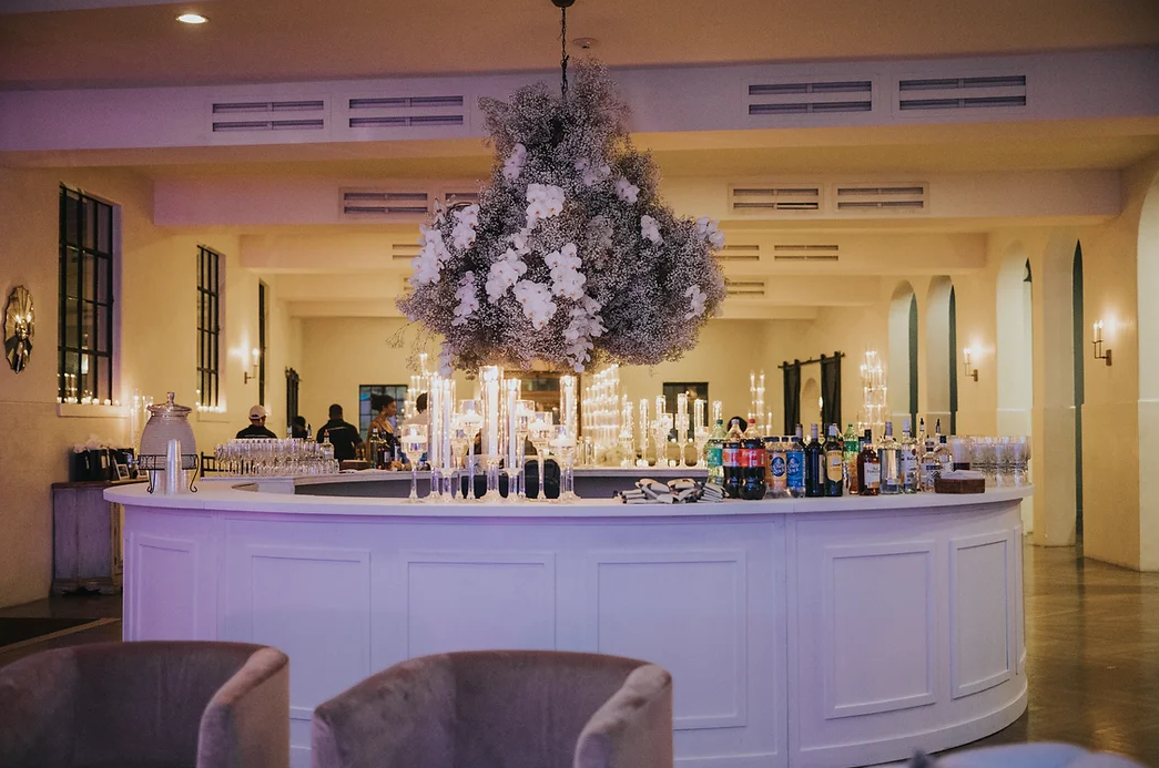 Elegant bar with floral centerpiece, surrounded by glassware and bottles, in a sophisticated event space with warm lighting.