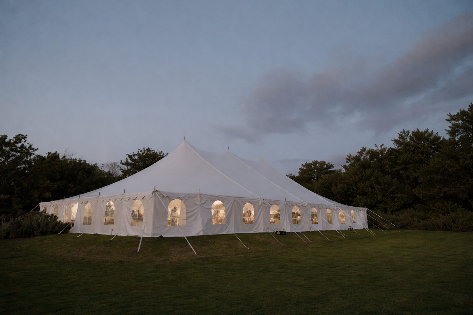 Large white event tent with illuminated interior, set up on a grassy area during dusk, surrounded by trees.