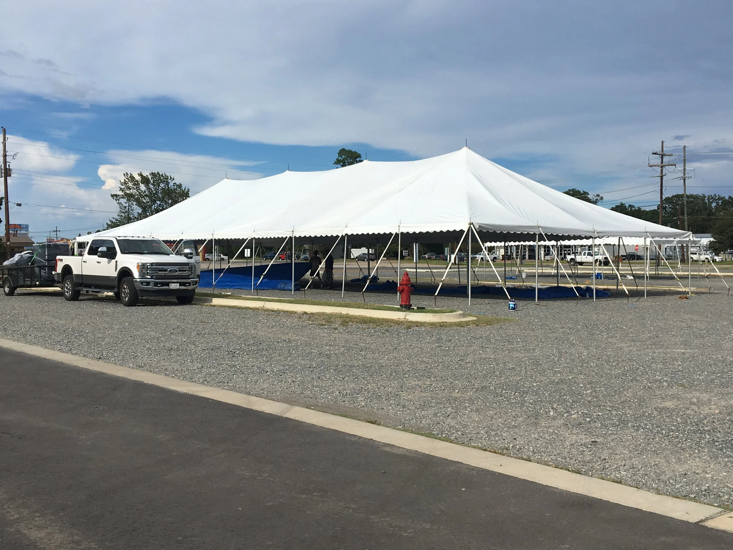 Large white event tent set up in a parking lot with a blue ground tarp underneath, parked vehicles nearby, and a partly cloudy sky overhead.