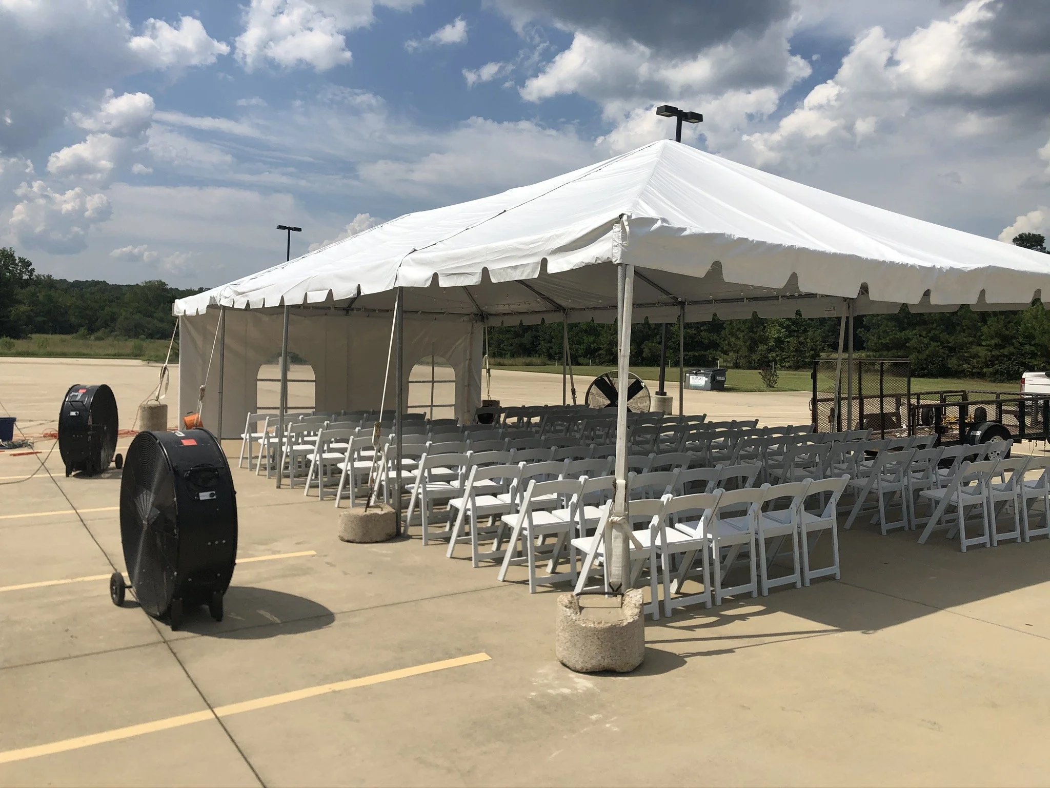 Setup of chairs under a large white event tent in a parking lot with fans localized around.