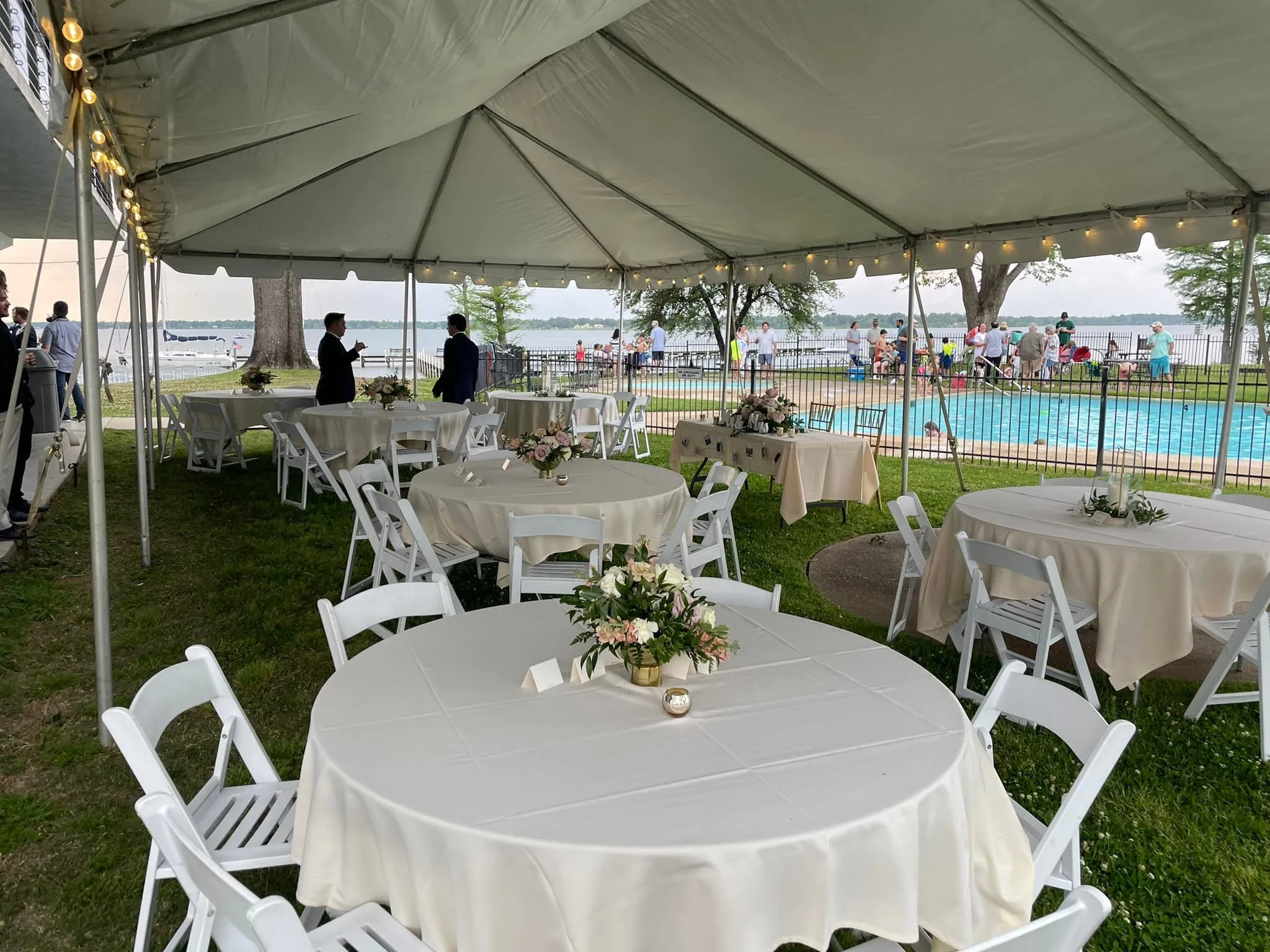 Under a large tent with string lights, several round tables with white tablecloths and floral centerpieces are set up on a grassy area for an outdoor event. In the background, near a swimming pool, people are gathered and enjoying the outdoor setting by a body of water, with boats docked nearby.