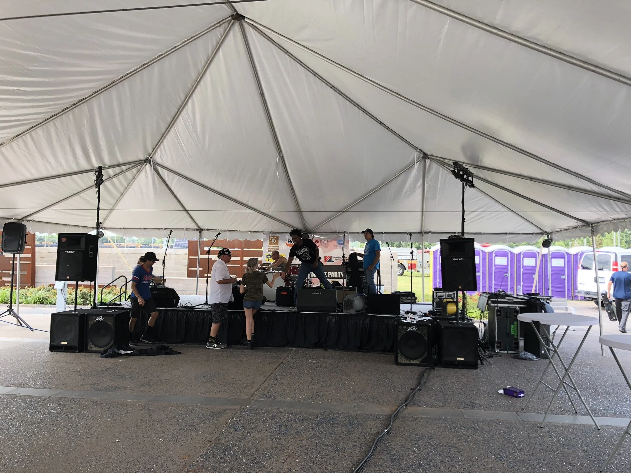 People setting up sound equipment on a stage under a large white tent during an outdoor event, with portable toilets and vehicles visible in the background.
