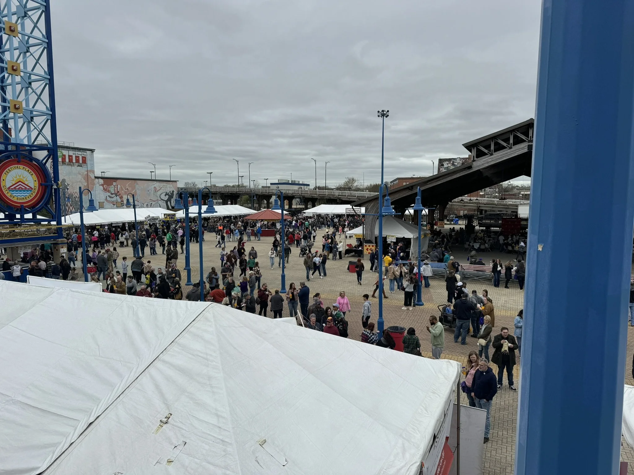 Crowd of people at an outdoor event with tents and a roller coaster in the background on an overcast day.