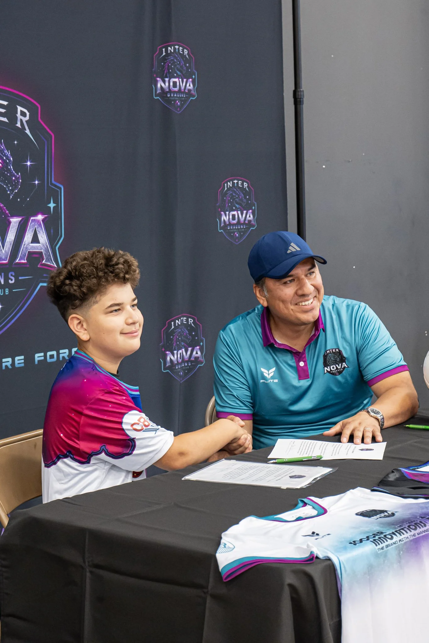 A young man and an older man are sitting at a table, shaking hands, with documents and pens in front of them. They are at a signing event for Inter Nova Dragons, as indicated by a banner with logos in the background and sports jerseys on the table.