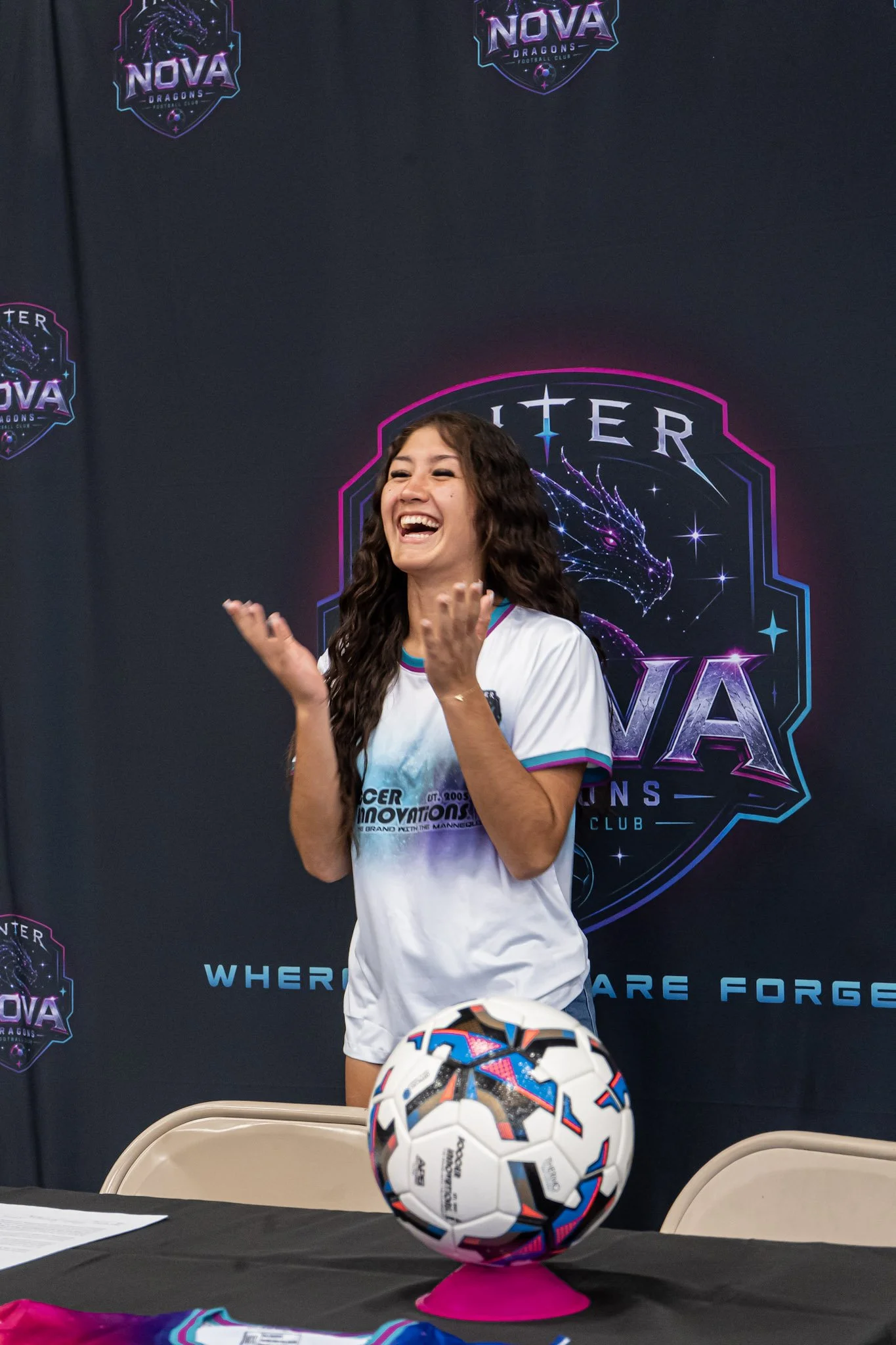 A woman with long curly hair smiling and laughing, wearing a white and blue soccer jersey, standing in front of a black backdrop with a colorful logo of Nova Dragons Football Club featuring a dragon and stars, with a soccer ball on a pink stand in the foreground on a table.