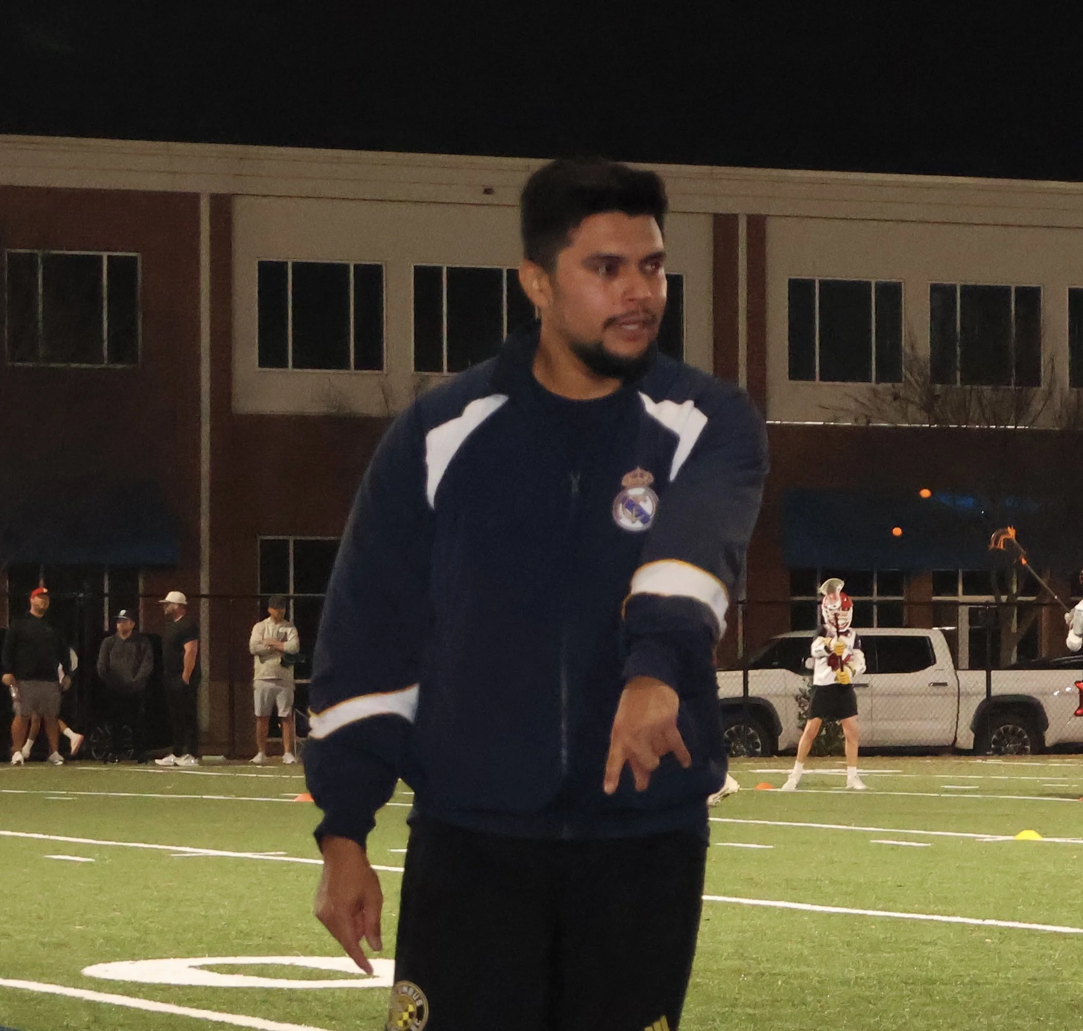 A young man with dark hair and a beard, wearing a dark sports jacket with a logo, standing on a sports field at night. In the background, people are gathered, and there is a building with large windows.