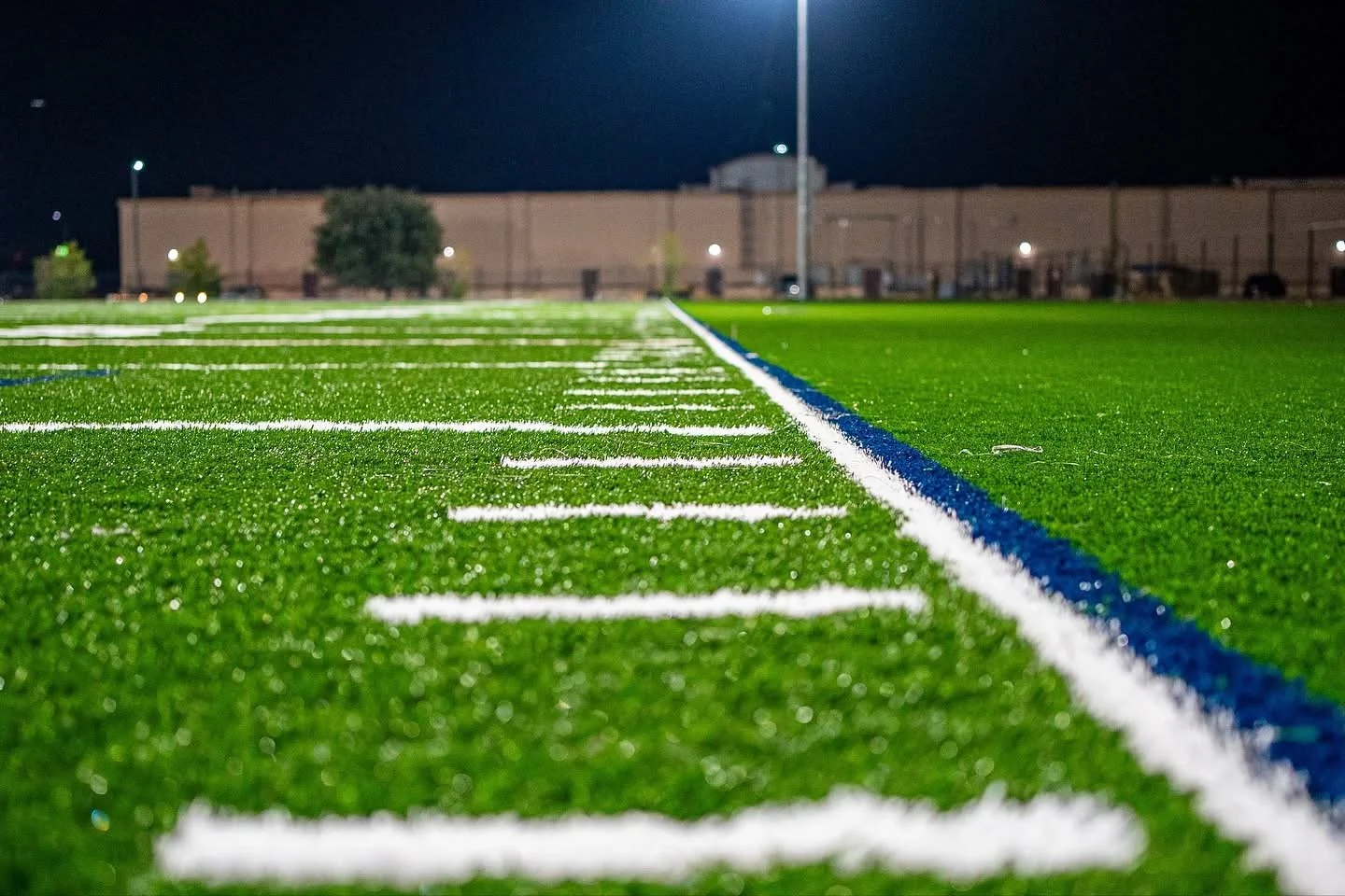 Close-up view of an empty synthetic football field at night, showing white yard lines and a blue sideline, with a building and trees in the background.