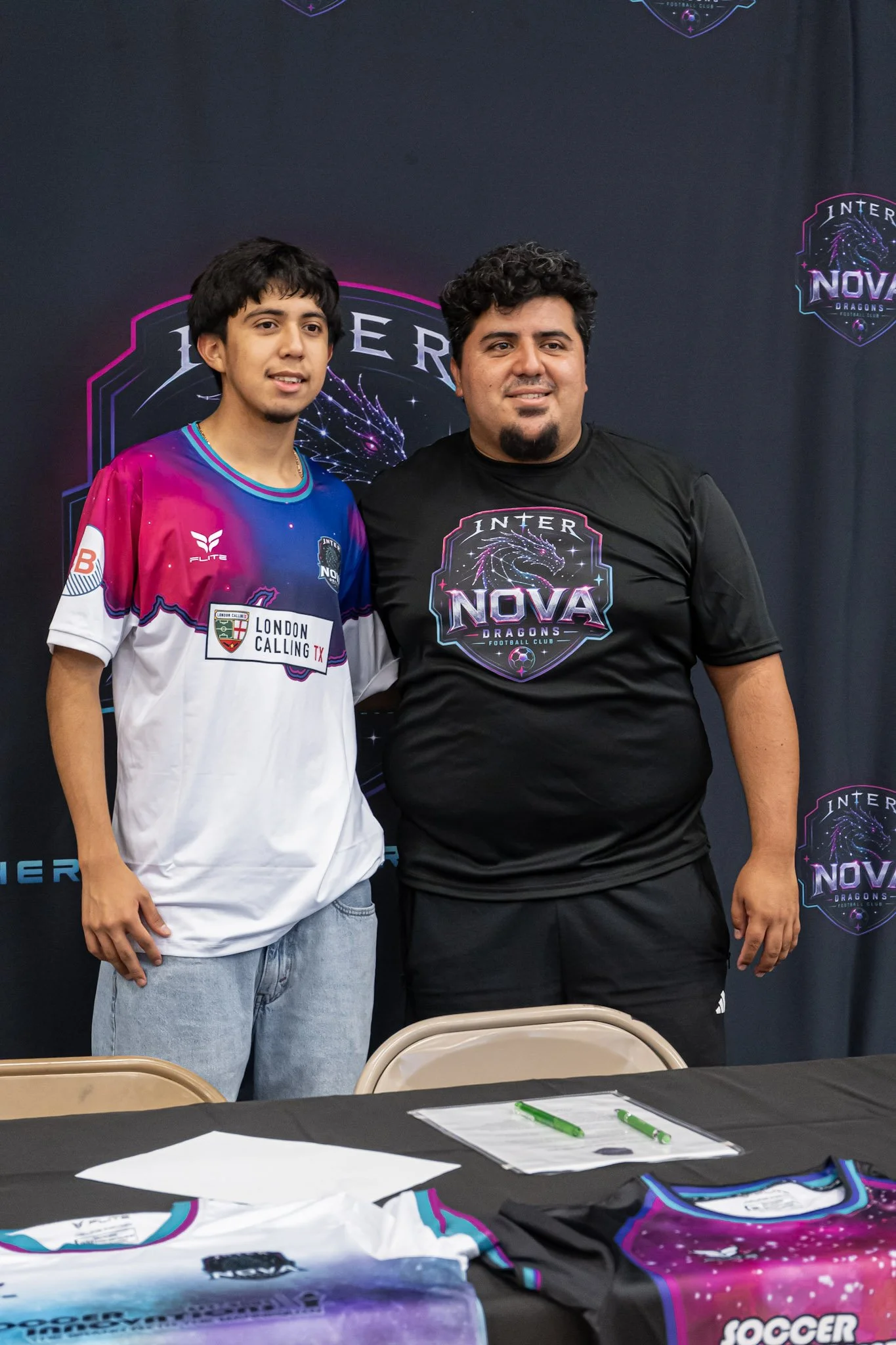 Two men stand side by side in front of a backdrop with the logo of Inter Nova Dragons esports team. The man on the left wears a colorful jersey with sponsor logos, and the man on the right wears a black t-shirt with the team logo. In the foreground, there are tables with papers, green pens, and a folded esports jersey.