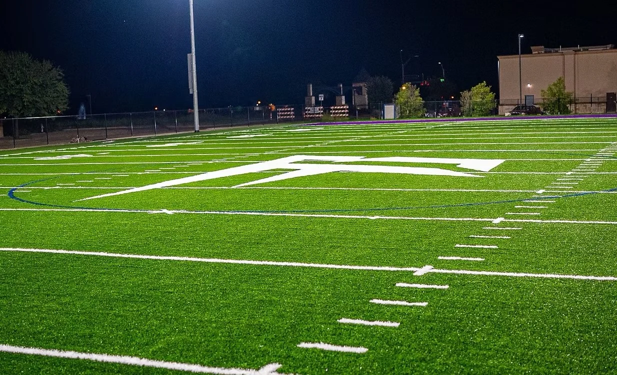 An illuminated sports field at night with bright green artificial turf and white yard lines.