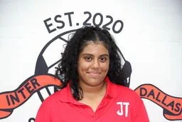 A young girl with dark curly hair wearing a red sports jersey standing in front of a white background with a logo that says 'EST. 2020' and includes the words 'INTER' and 'DALLAS'.