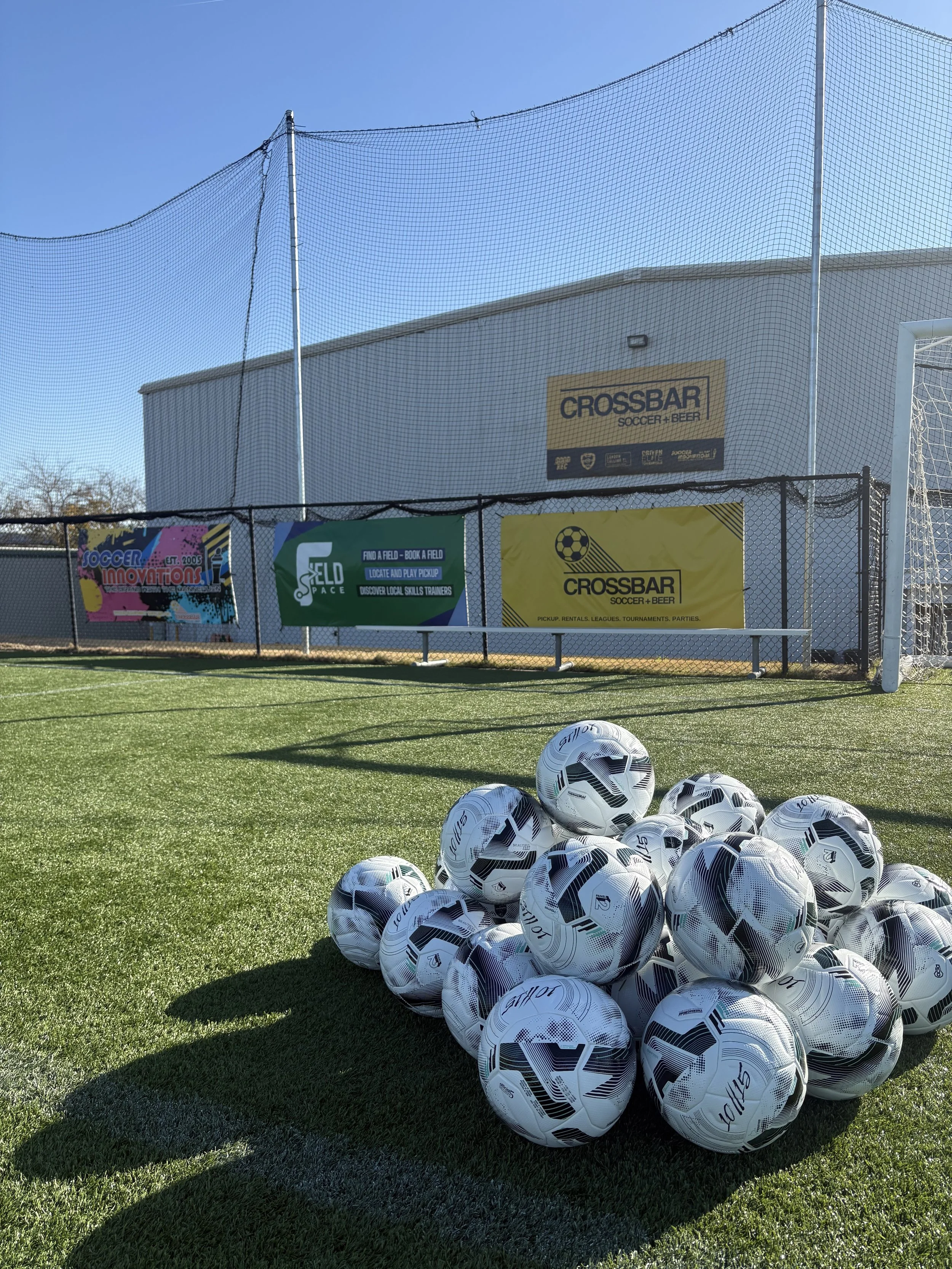 A pile of white and black soccer balls on green artificial turf at an outdoor soccer field, with a goal post and net, advertising banners on a fence, and a building in the background.