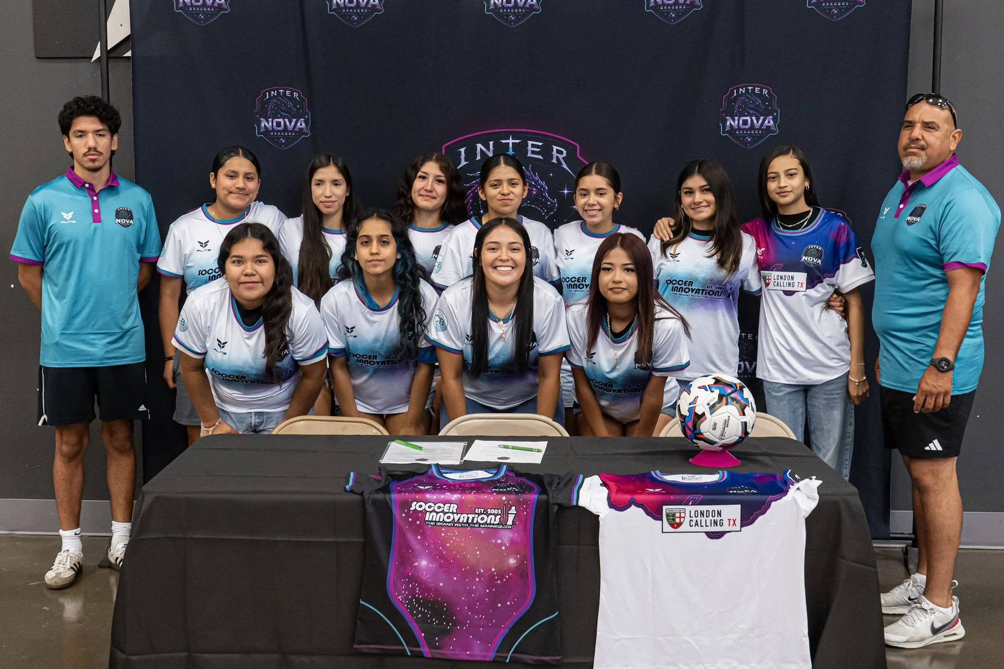 Group of young female soccer players posing with two coaches or staff members in front of a dark backdrop with the 'Inter Nova Dragons' logo. There are soccer jerseys, a signed soccer ball, and promotional items on the table in front of them.