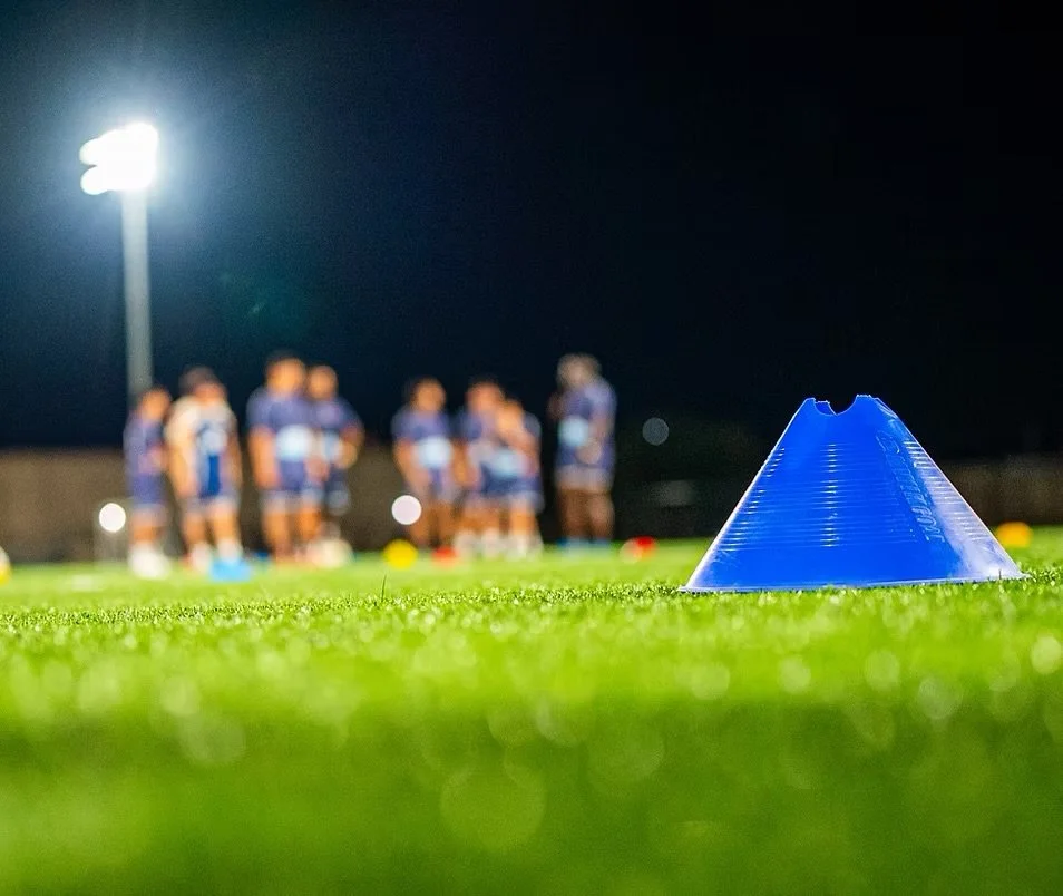 A soccer cone on a grassy field with a group of players blurred in the background, illuminated by stadium lights at night.