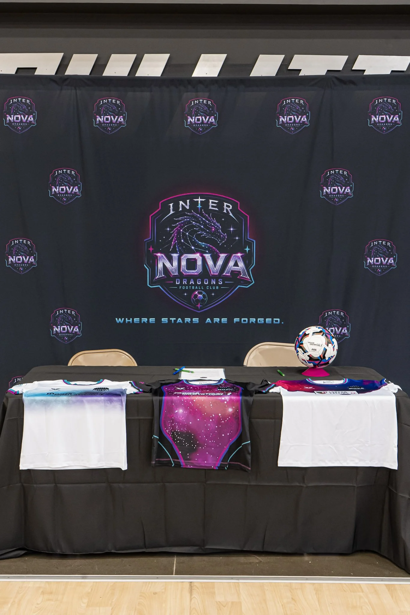 A display table with various merchandise, including a framed shirt, a pillow, and a scarf, set against a backdrop with the Inter Nova Dragons logo and the slogan 'Where stars are forged'.