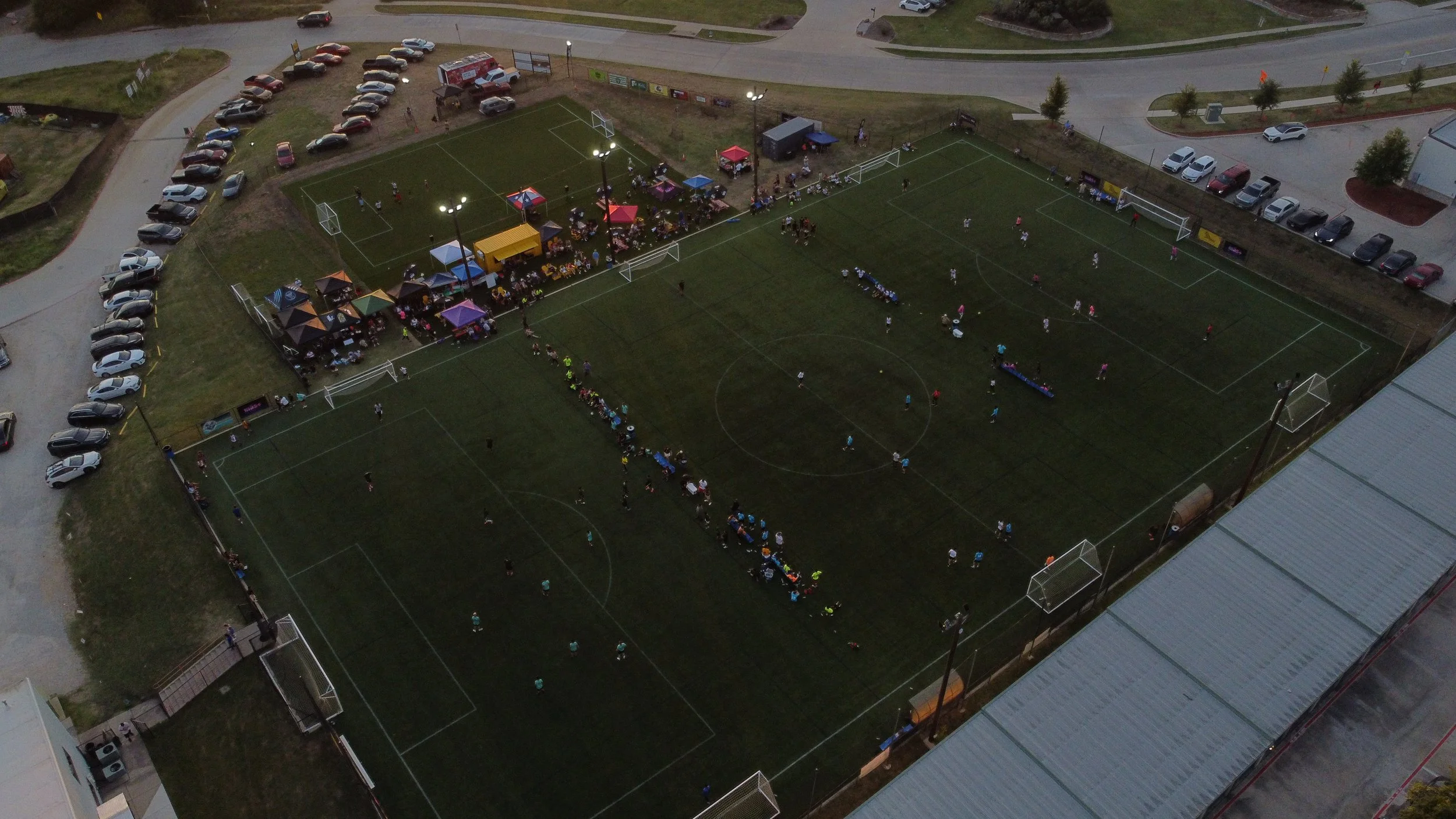 Aerial view of a soccer field with players and spectators, surrounded by parking lots and a small gathering area with tents and tables.