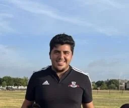 A smiling man with dark hair wearing a black sports shirt standing outdoors on a grassy field.