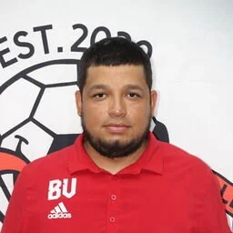 Portrait of a man with short dark hair and a beard wearing a red polo shirt with 'BU' and Adidas logo, standing in front of a white wall with a circular logo and text.