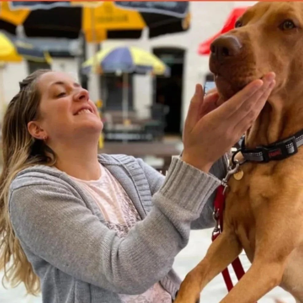 A woman with long blonde hair and a light gray sweater is playing with a large brown dog, holding its face gently. They are in an outdoor setting with colorful umbrellas and outdoor furniture in the background.