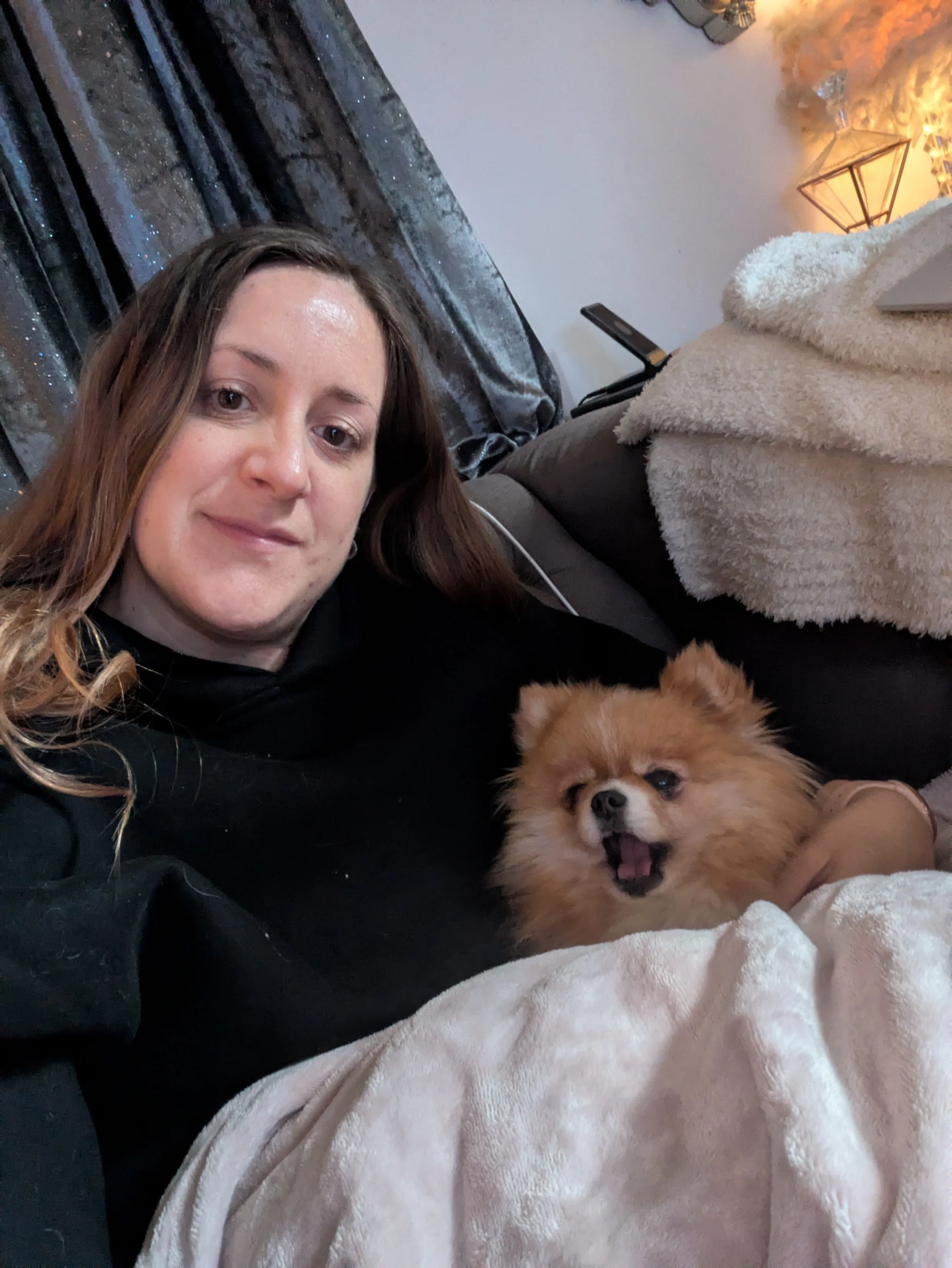 A woman with long brown hair lying on a couch next to a small fluffy dog with tan fur. The woman is smiling lightly, and the dog appears to be barking or yawning with its mouth open. There are blankets and pillows around them, and a lamp with warm li