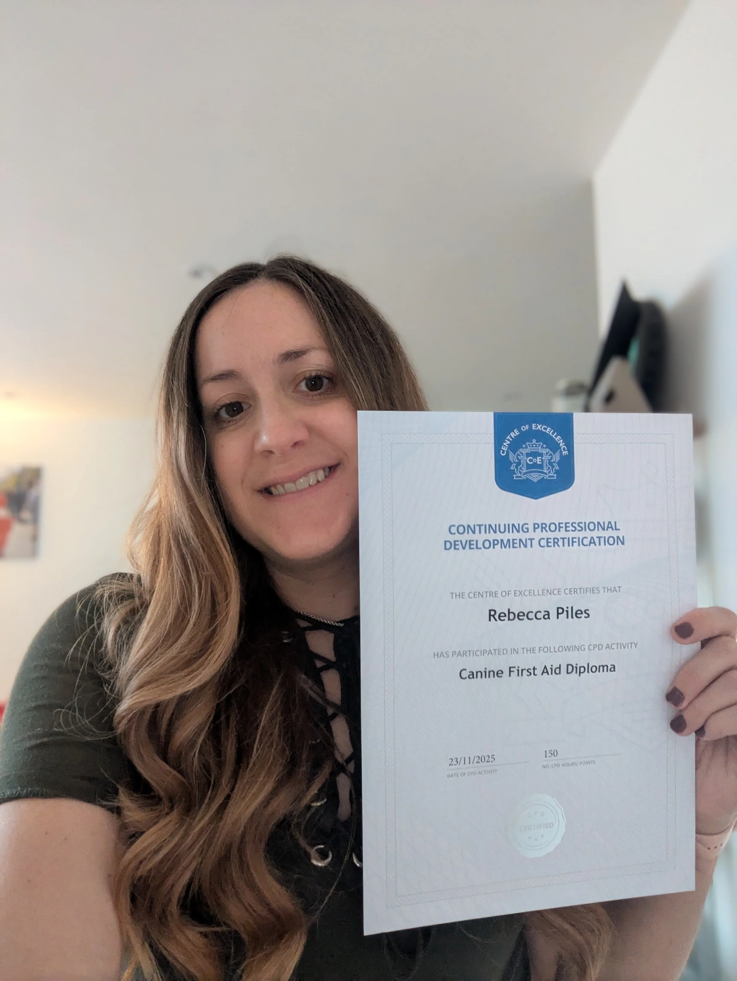 A woman with long wavy brown hair holding a certification for a Canine First Aid Diploma, smiling at the camera inside a room with light-colored walls.