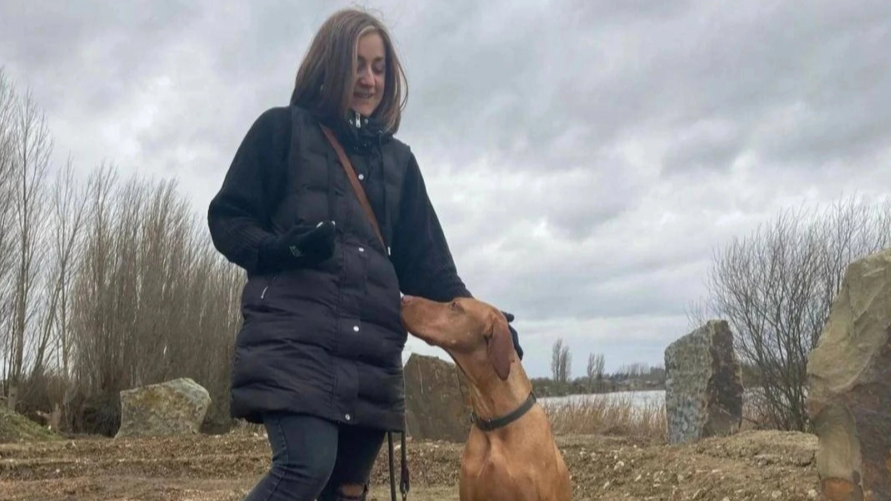 A woman in a black jacket interacts with a brown dog near a lakeside, overcast sky, with leafless trees and rocks in the background.