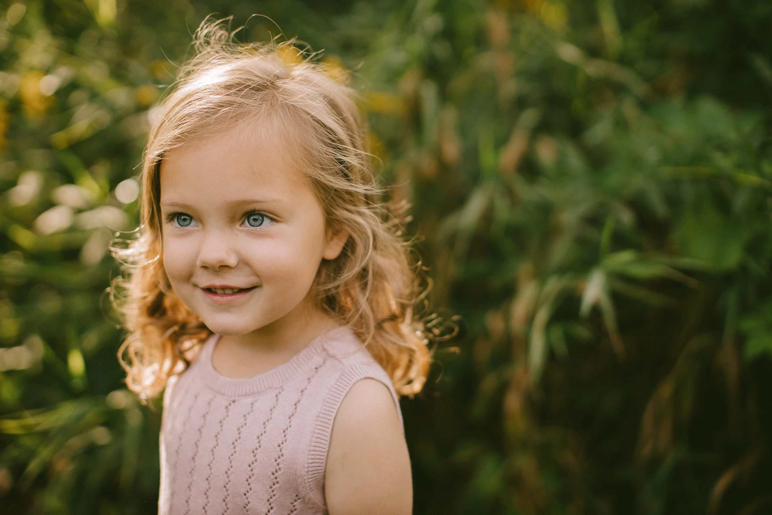 A young girl with curly blonde hair and blue eyes smiling outdoors in a green, leafy environment.
