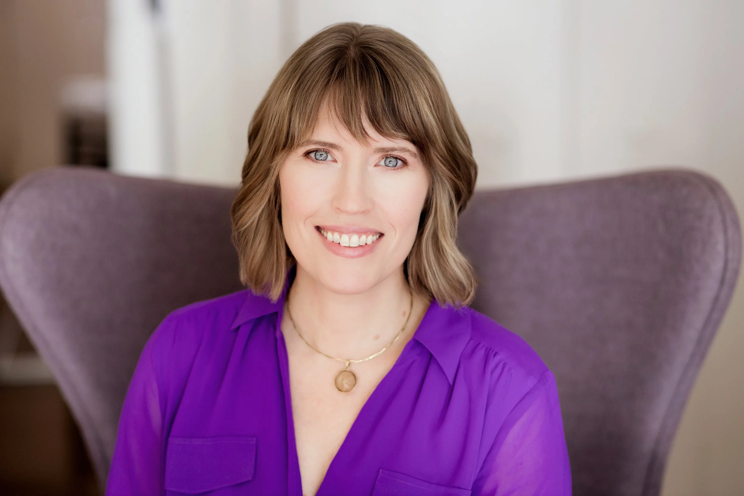 Portrait of a woman with shoulder-length light brown hair, blue eyes, and a bright smile, sitting on a purple chair, wearing a purple blouse and a gold necklace with a round pendant. Headshot photography session in Evanston, IL.