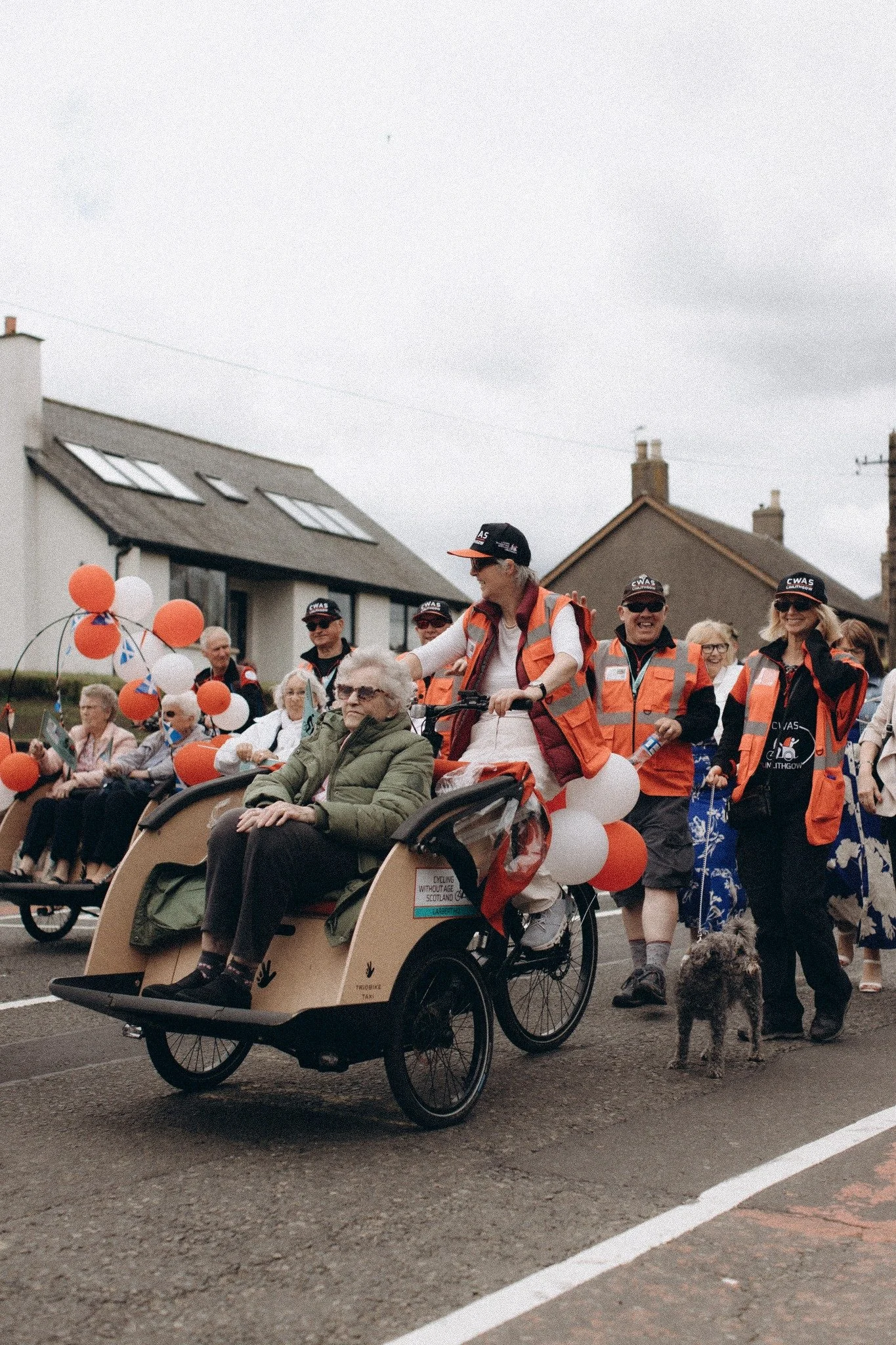 A parade featuring elderly individuals, with some in wheelchairs, being guided by volunteers wearing orange safety vests and hats, some of whom are riding bicycles or walking dogs. The participants are decorated with orange and white balloons, and th