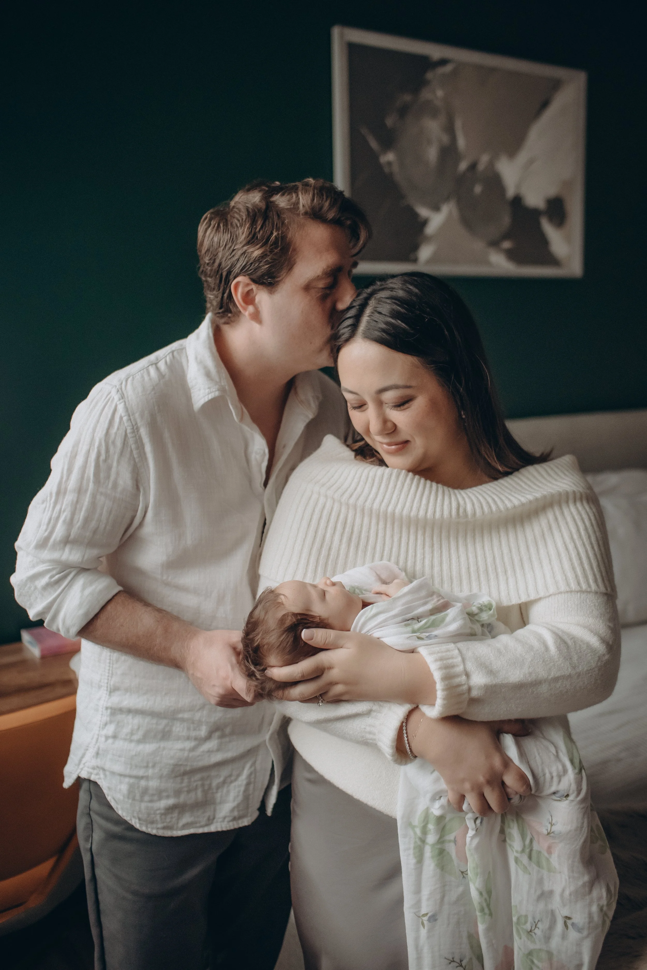 A couple holding their newborn baby in a bedroom, with the father kissing the mother's forehead. Lifestyle newborn session shot in Chicago IL.