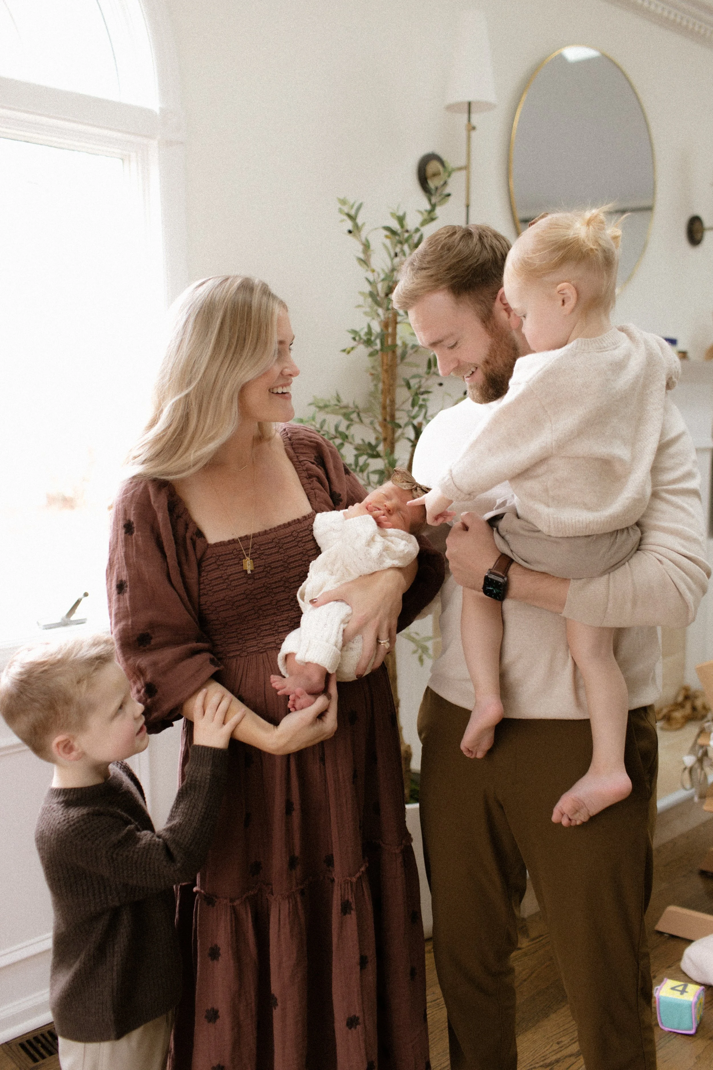 A family of five, including a woman, a man, one young boy and two babies, stands indoors near a window with a mirror and plants behind them. The woman holds a newborn baby, and the man holds a toddler reaching towards the baby's face. The scene appea