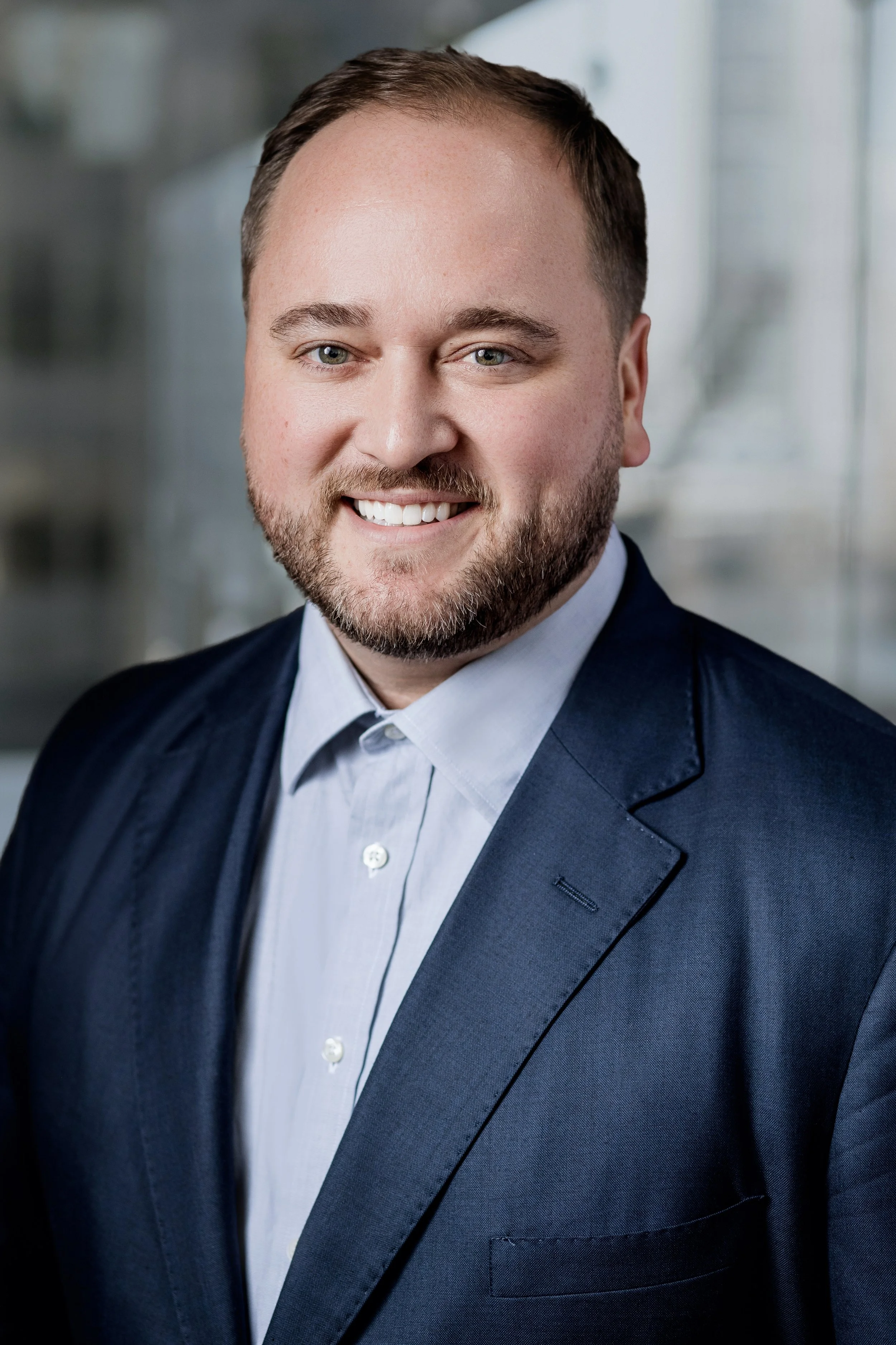 A professional headshot of a man with a beard and short brown hair, wearing a navy blue blazer and white dress shirt, standing in front of a blurred office background. Headshot photography session in Evanston, IL.