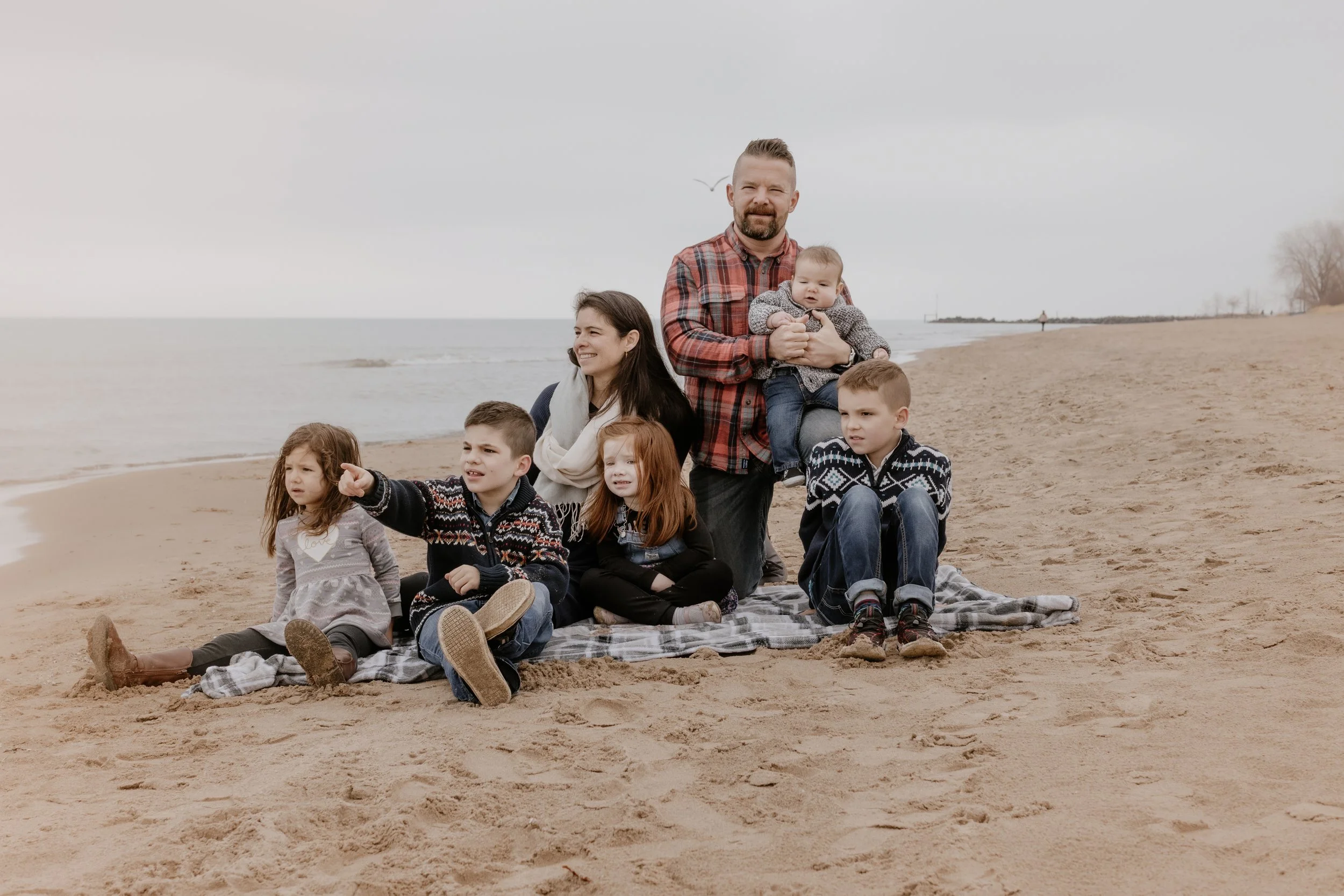 A family of seven sitting on a blanket on a sandy beach, with the ocean in the background. The father is holding a baby, while the others, including two girls and two boys, are seated around them. The family is dressed in casual, warm clothing suitab
