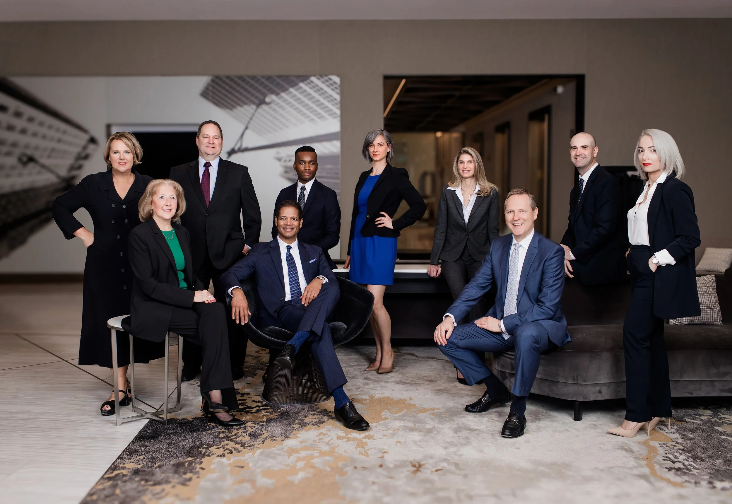 A diverse group of ten professionals, five women and five men, posing in a modern office with a cityscape mural on the wall behind them. They are dressed in business attire, some standing and some sitting, smiling confidently.