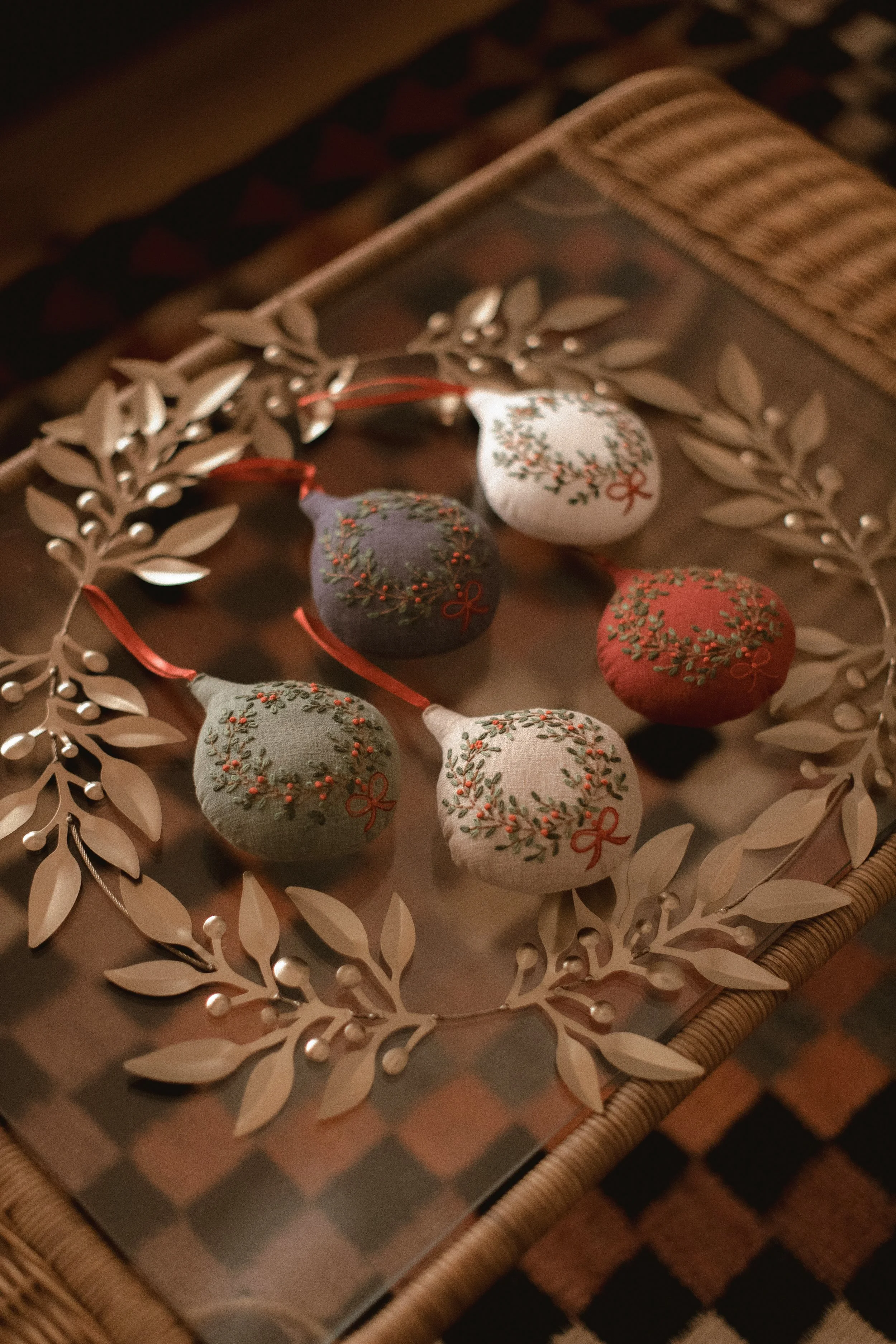 Five fabric Christmas ornaments shaped like pears with embroidered holly and red bows, displayed on a decorative mirror with a metallic leafy border. Product Photography Shoot Evanston, IL.