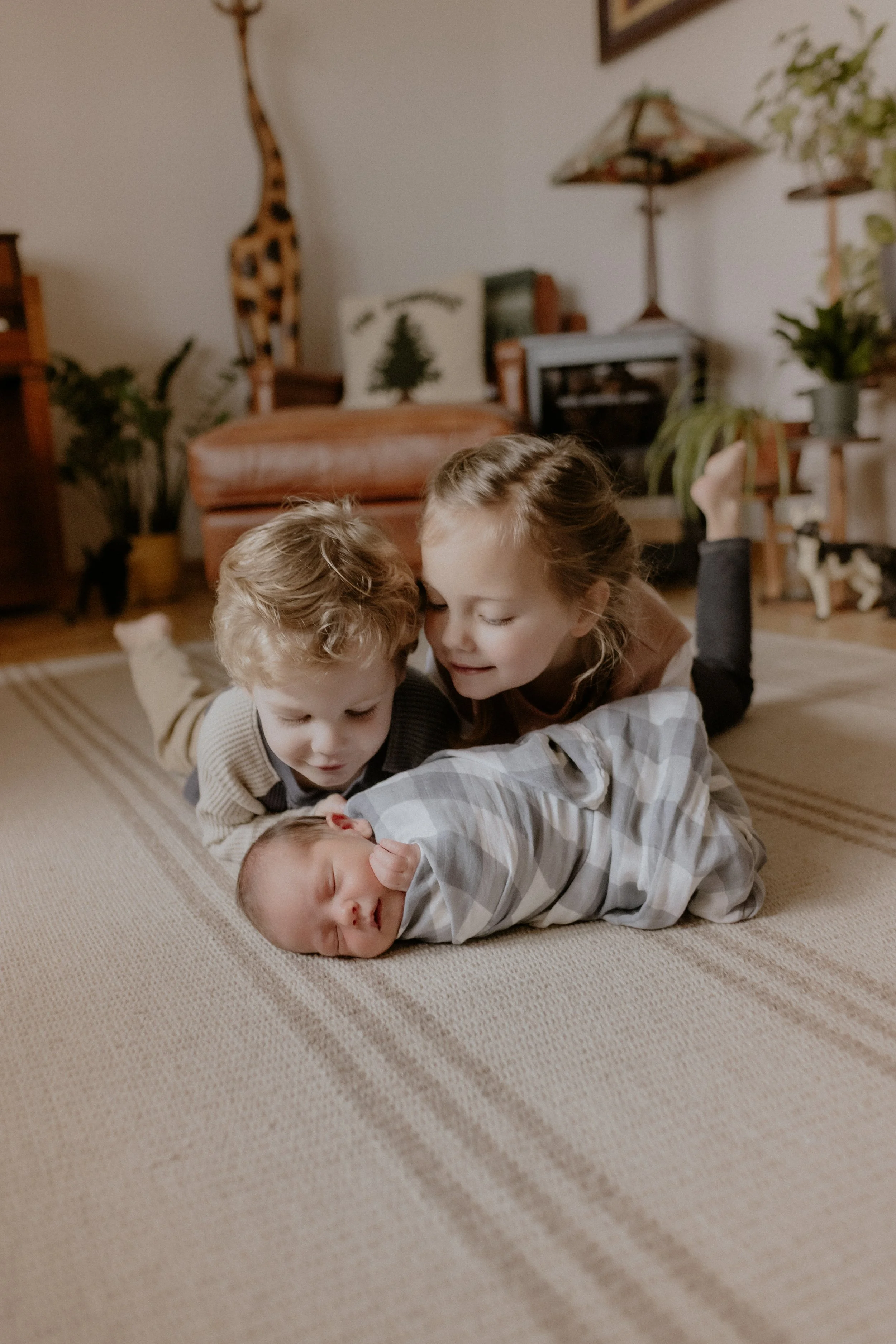 Three children lying on a beige carpet, with a newborn baby sleeping in the middle. The older children are leaning over the baby, smiling and looking at them. Lifestyle Newborn Photography Session Chicago, IL.
